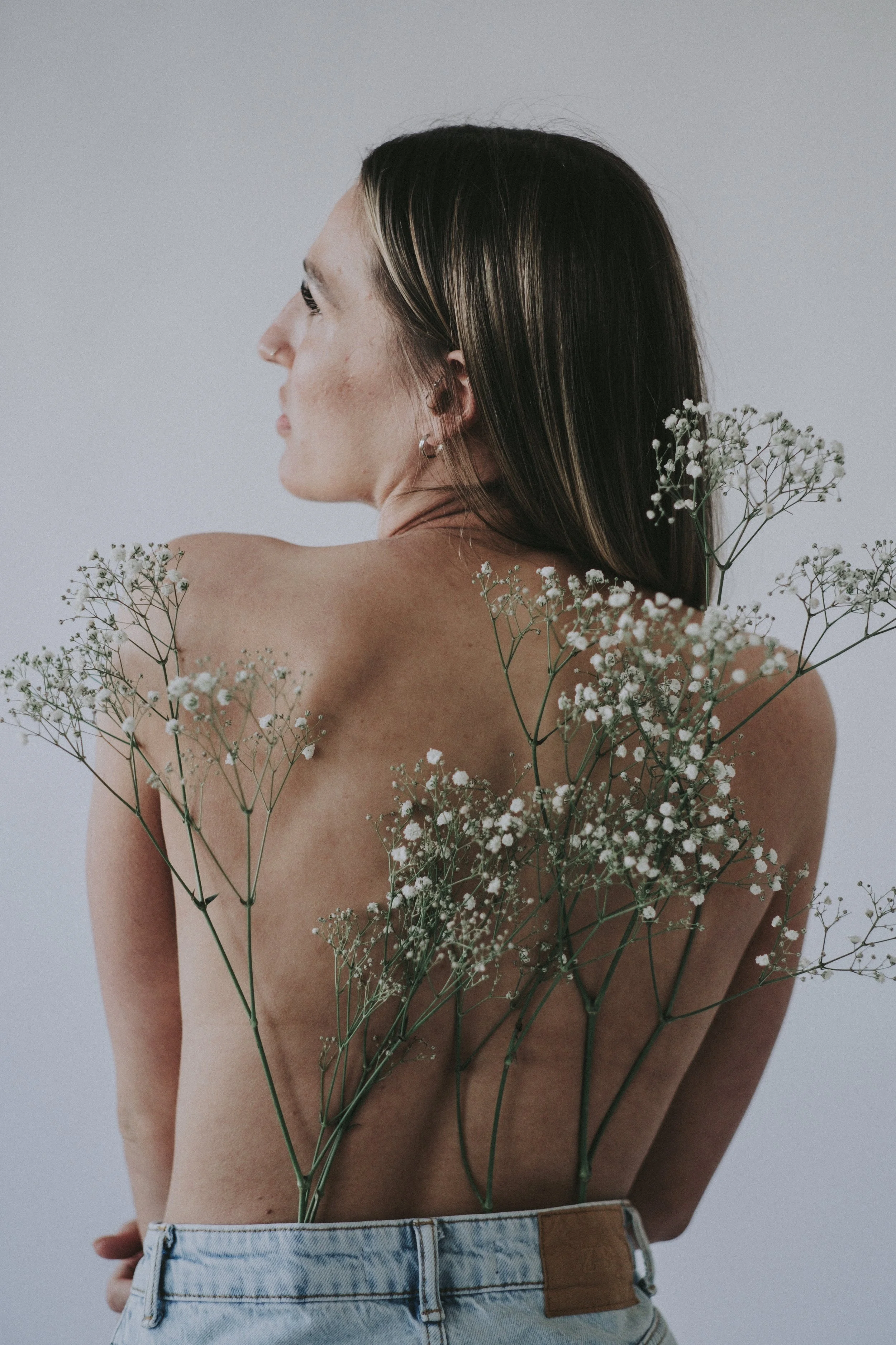 A woman with long, dark hair and earrings, seen in profile, with white flowers resting on her shoulders and back, wearing jeans, against a plain light background.