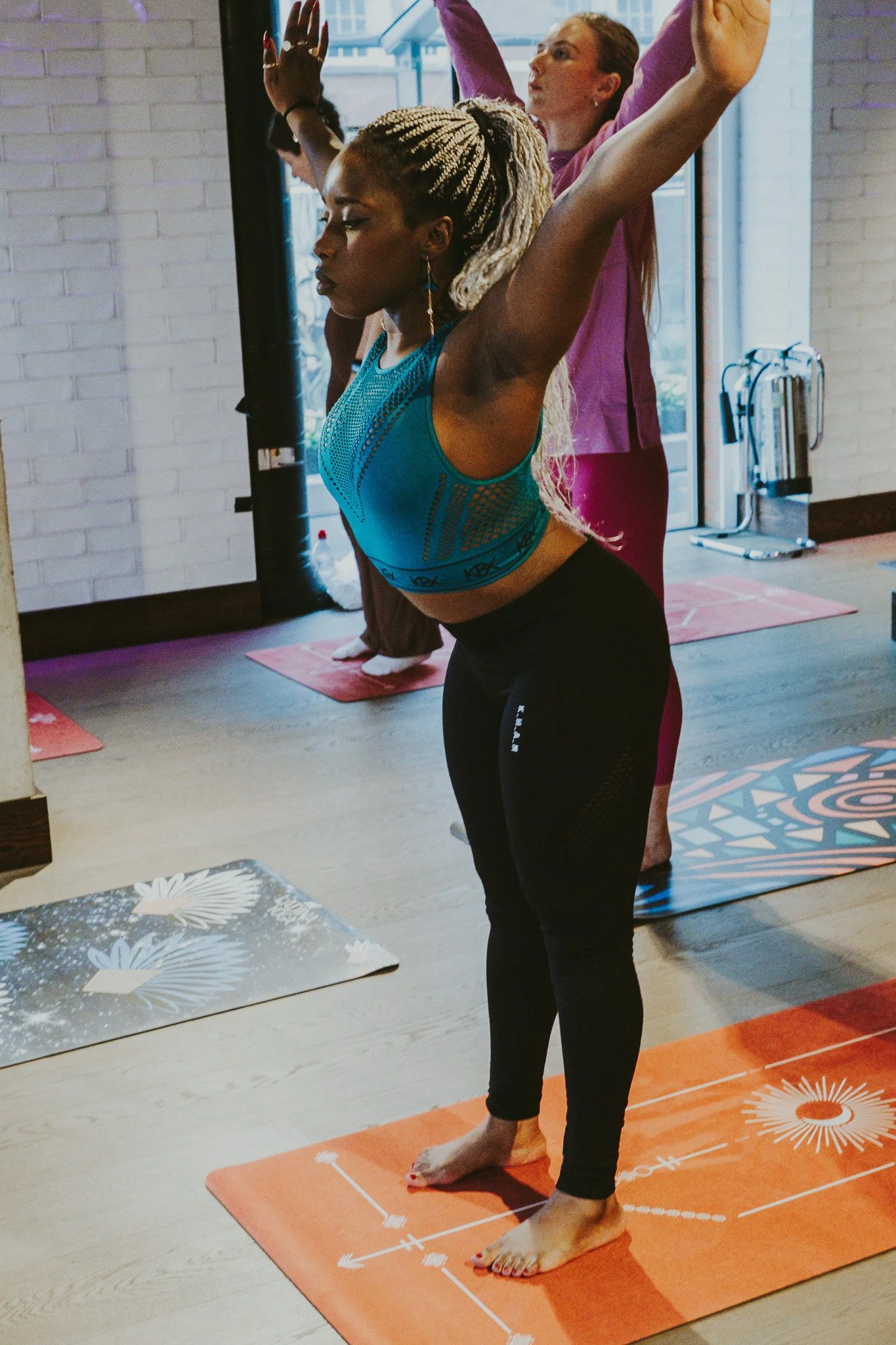 Four women practicing yoga, standing on colorful yoga mats inside a room with brick walls and large windows, with their arms raised above their heads.