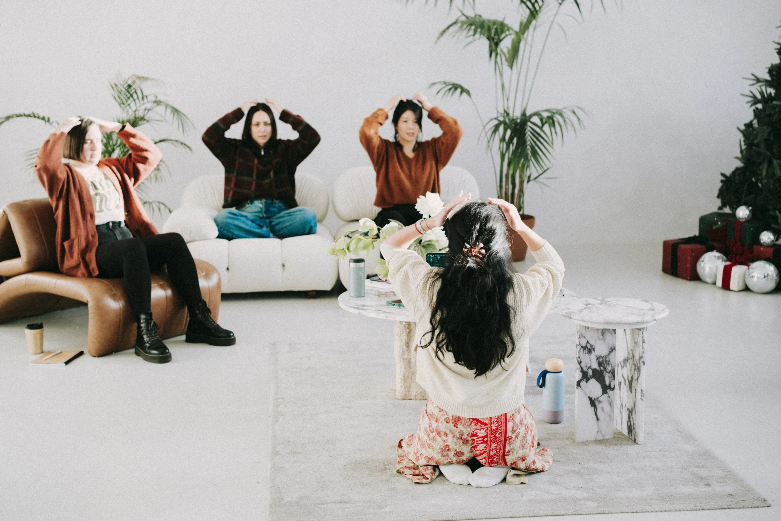 Four women participating in a yoga or meditation session indoors, with a Christmas tree and presents in the background.