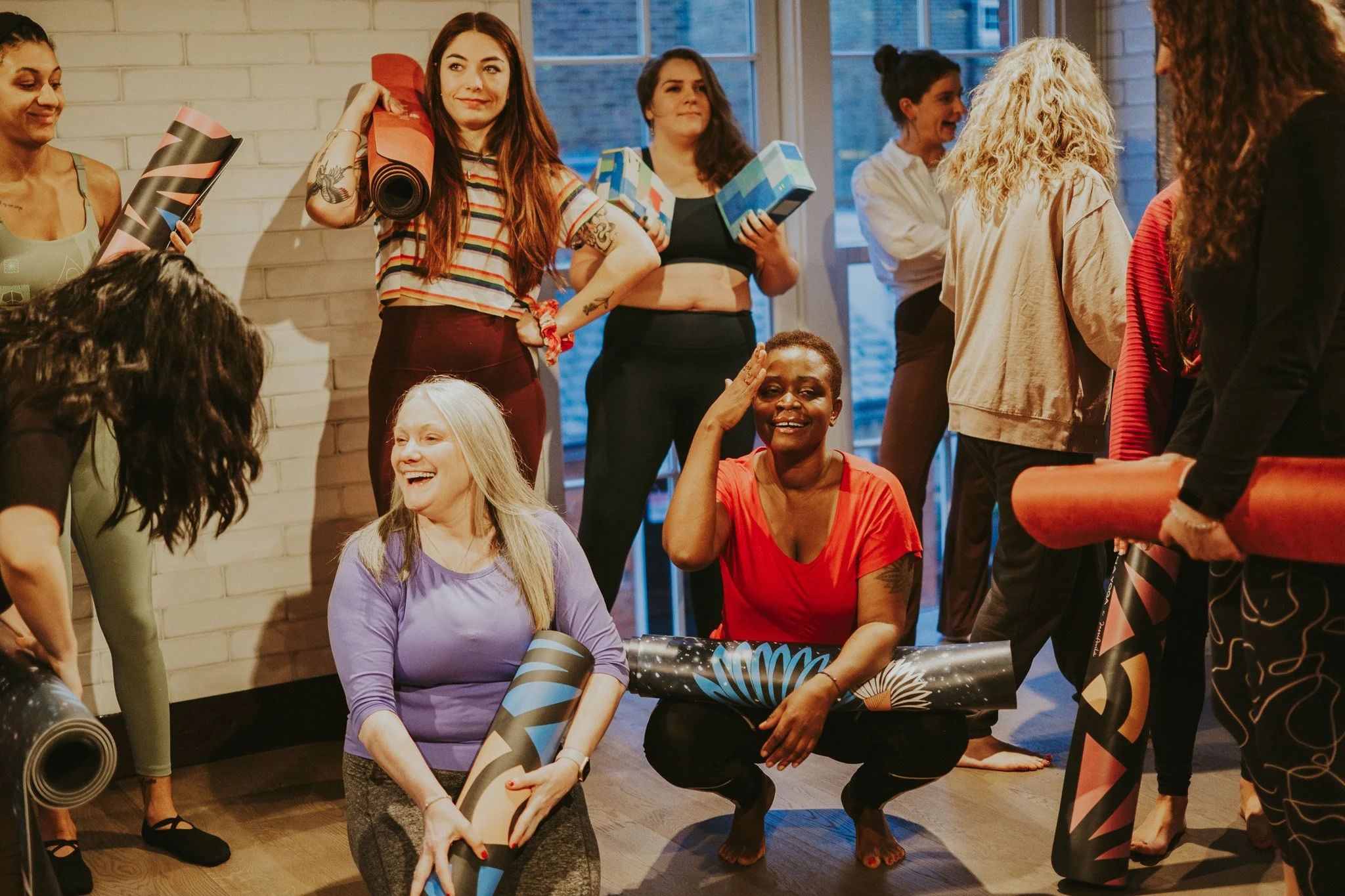 Group of women at a fitness or yoga class, some holding rolled yoga mats, some sitting and others standing, in a bright indoor space.