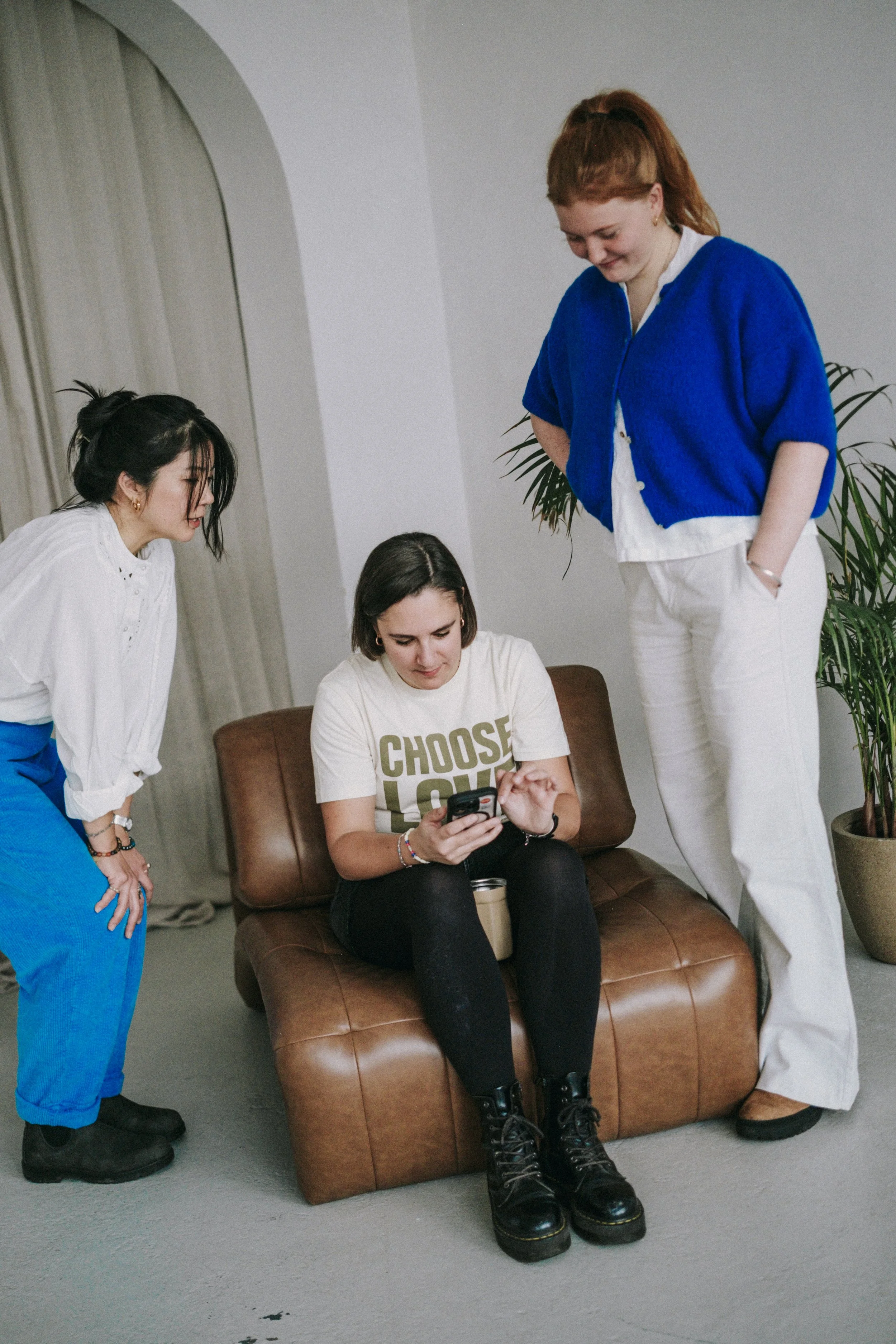 Three young women in casual clothing are gathered around, looking at a phone held by the woman sitting on a brown leather couch. The woman on the left is leaning forward, wearing a white shirt and blue pants. The woman on the right is standing, wearing a blue cardigan and white pants. The woman in the middle is seated, wearing a white t-shirt with the words 'CHOOSE LOVE' and black pants, focused on her phone.