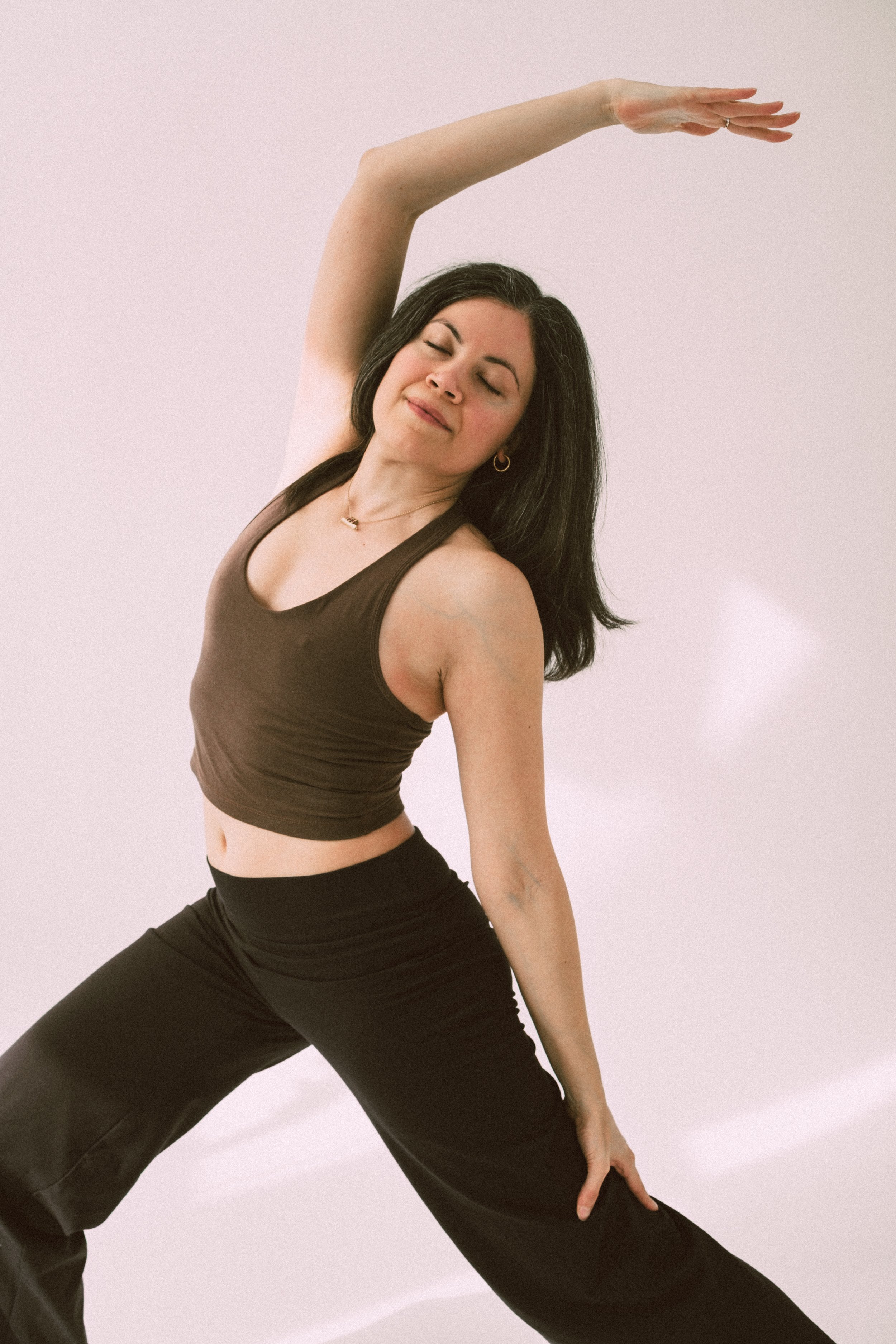 A woman with dark hair, wearing a brown tank top and black pants, practicing yoga in a wide stance with one arm extended overhead and the other resting on her thigh against a plain light pink background.