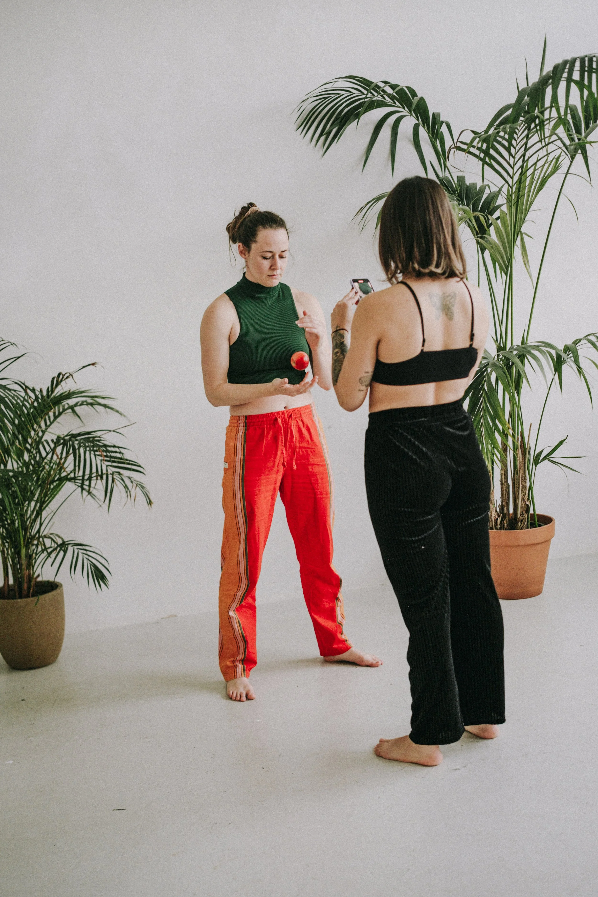Two women are standing barefoot on a white floor in a minimalist room with potted plants. One woman with dark hair in a messy bun, wearing a green sleeveless turtleneck and red striped pants, is holding a red apple. The other woman with dark, shoulder-length hair, wearing a black spaghetti strap top and black pants, is taking a photo with her phone, revealing a butterfly tattoo on her back.