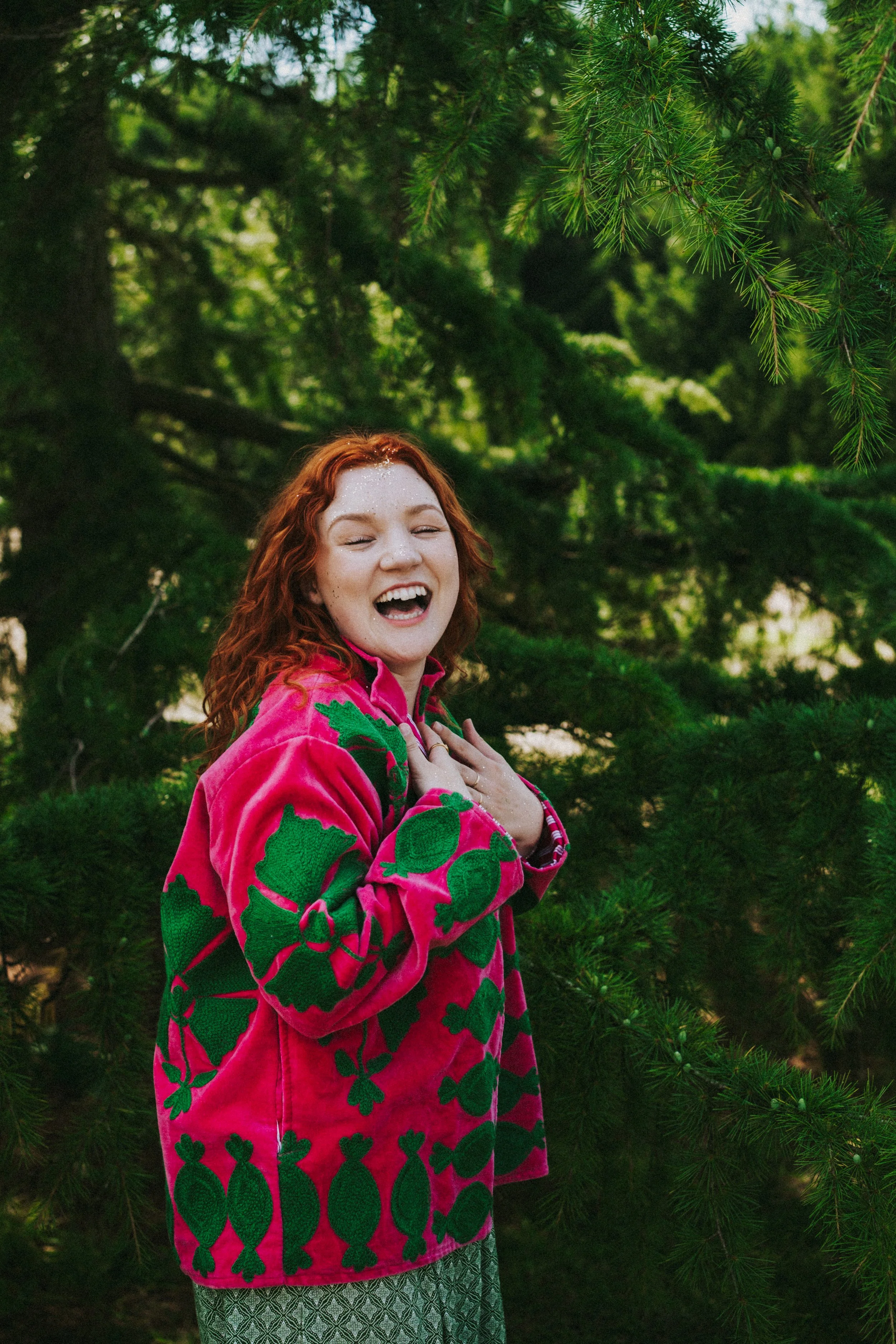 A joyful woman with long red hair, wearing a bright pink sweater with green leaf patterns, is standing outdoors in front of lush green evergreen trees, smiling and holding her hands to her chest.