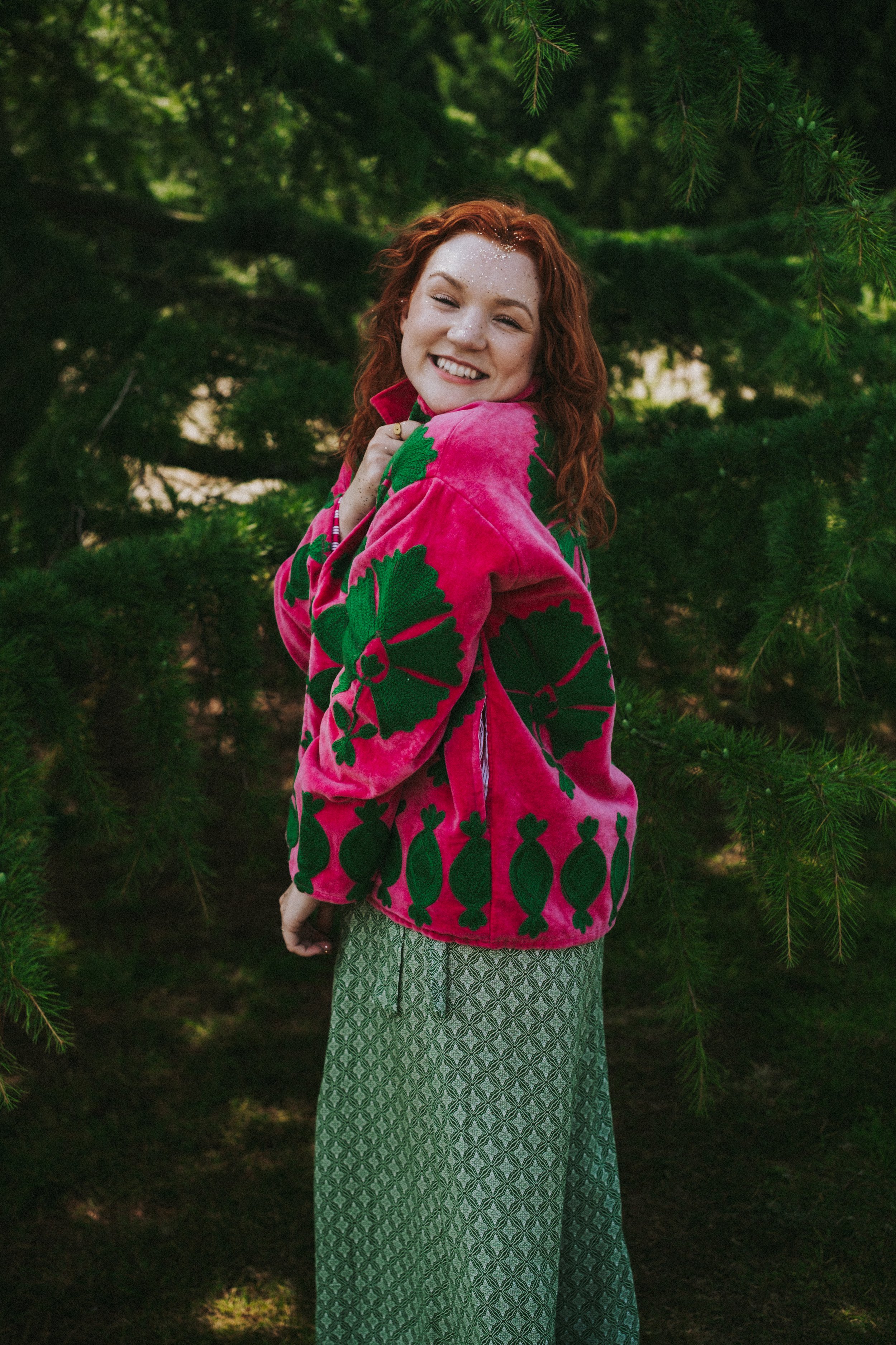 A woman with curly red hair smiling, standing amidst green pine trees. She is wearing a pink jacket with green leaf patterns and patterned green wide-leg pants.