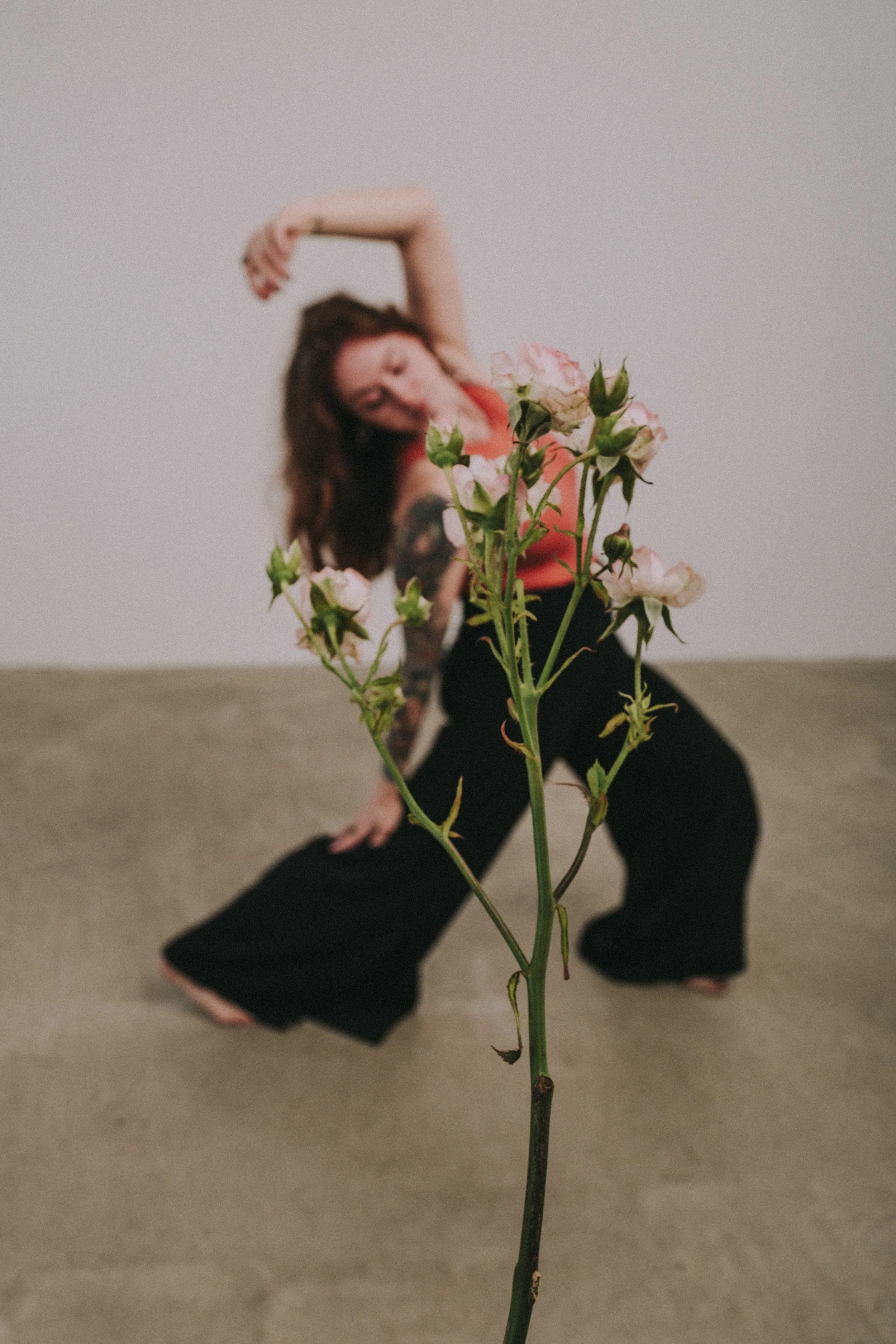 A woman with tattoos performing a dance pose in front of a plain wall, with a pink flower in the foreground.