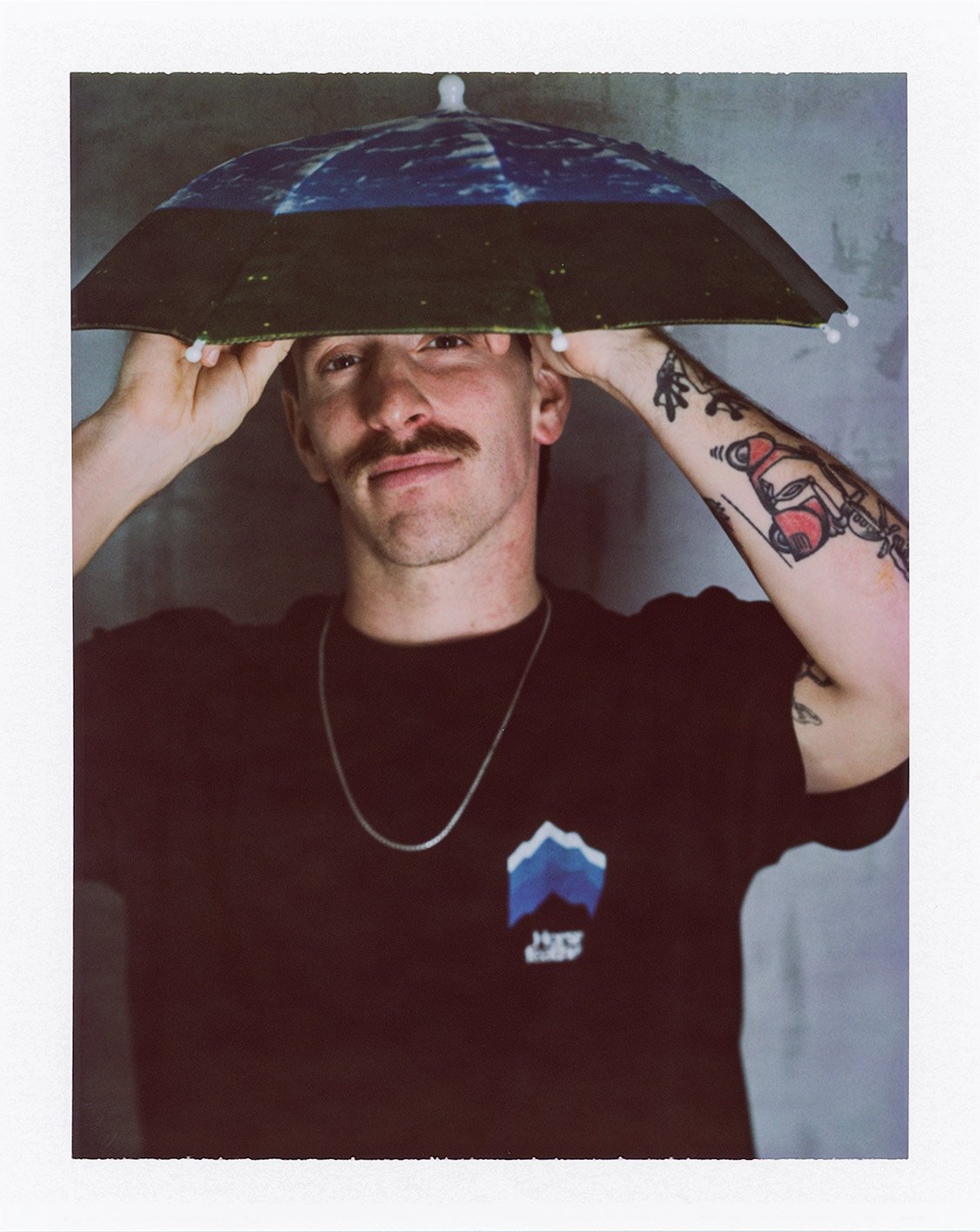 Mann mit Schnurrbart und Tätowierungen hält einen großen Regenschirm über dem Kopf und steht vor einem neutralen Hintergrund.
