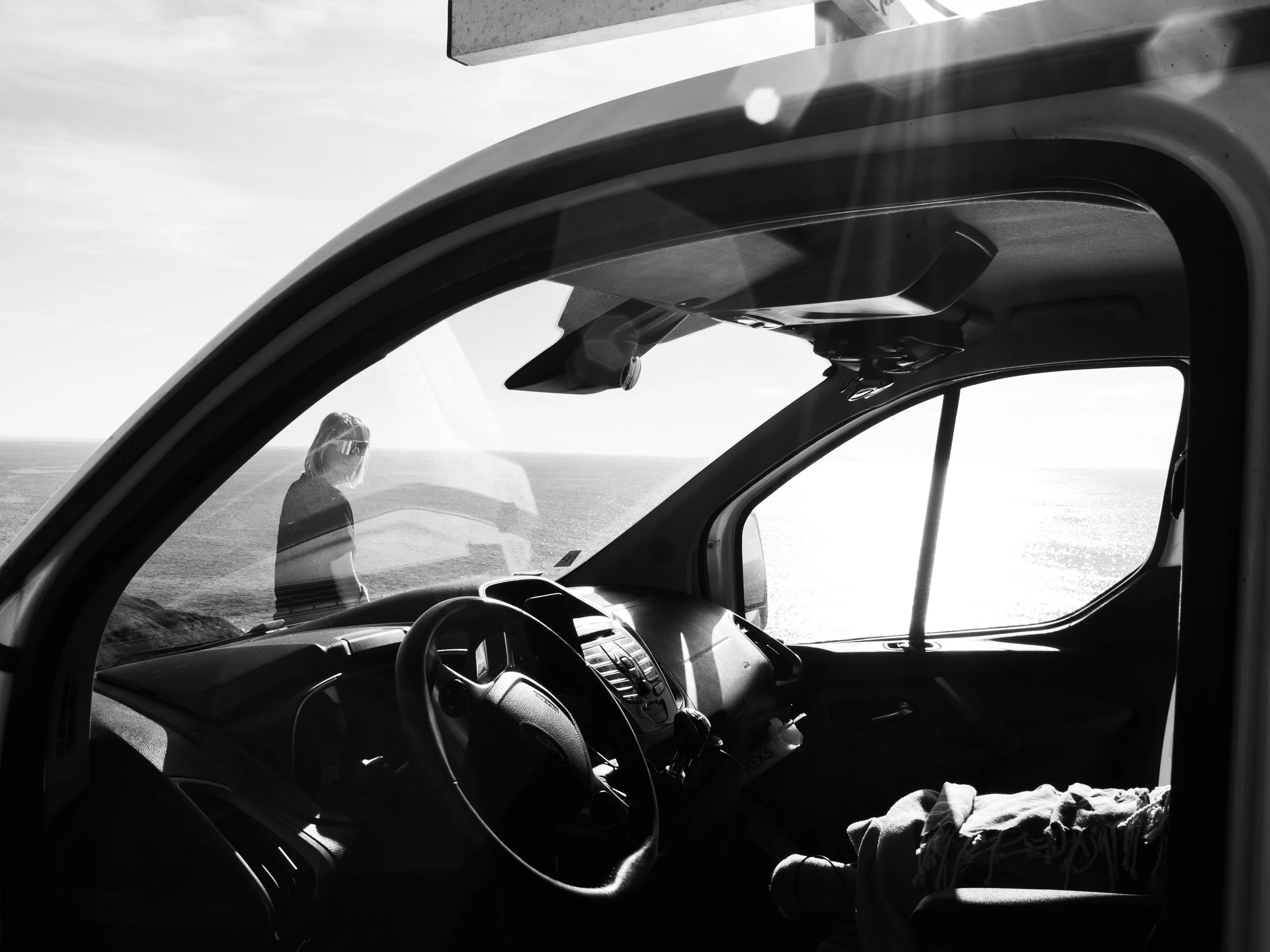 Blick aus dem Inneren eines Fahrzeugs auf eine Person, die draußen nahe dem Fenster steht und bei Sonnenuntergang auf das Meer blickt.
