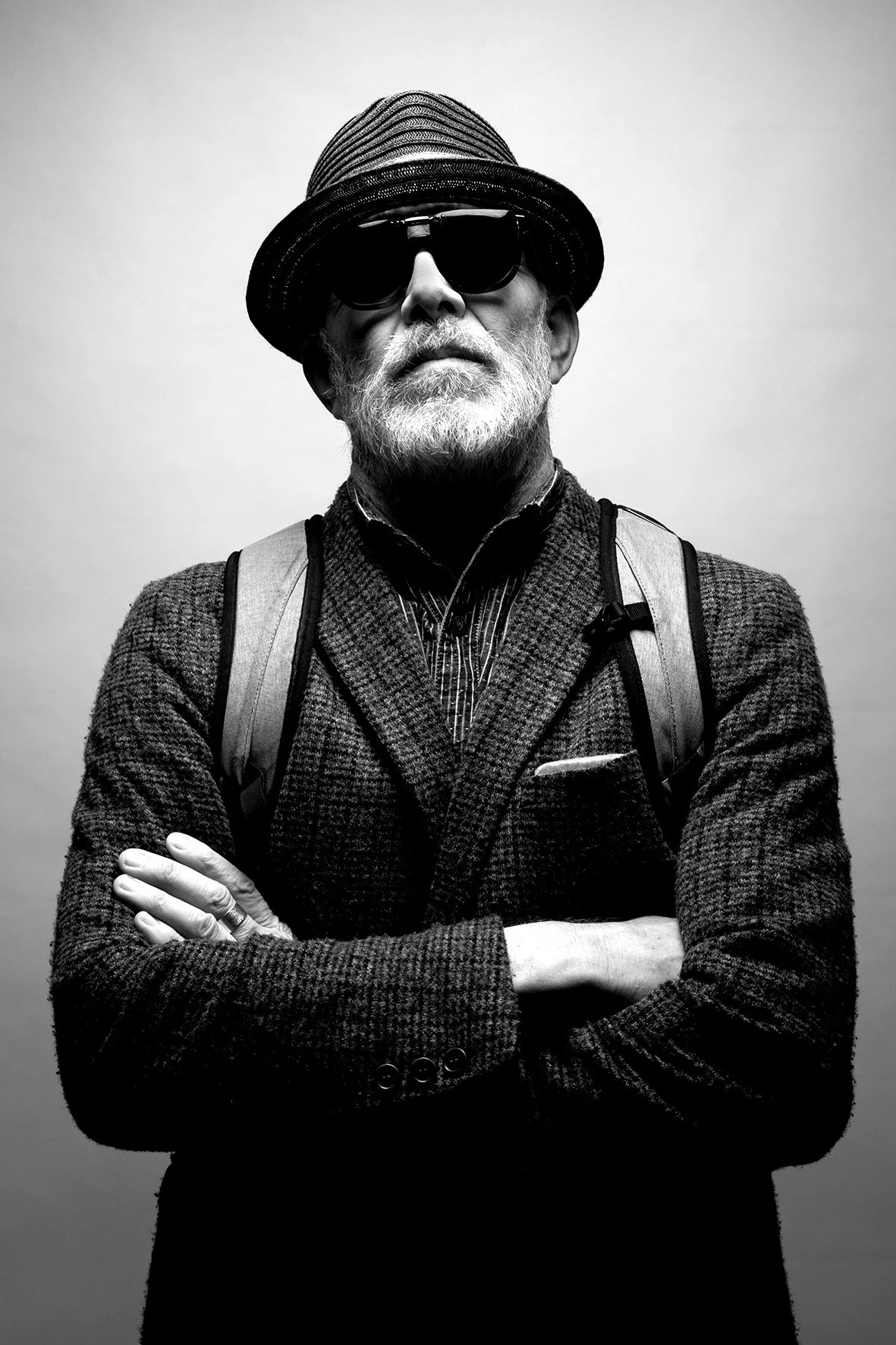 Selbstbewusster älterer Mann mit weißen Haaren und Bart, trägt Hut, Sonnenbrille und Jacke sowie einen Rucksack, steht mit verschränkten Armen vor einem neutralen Hintergrund.
