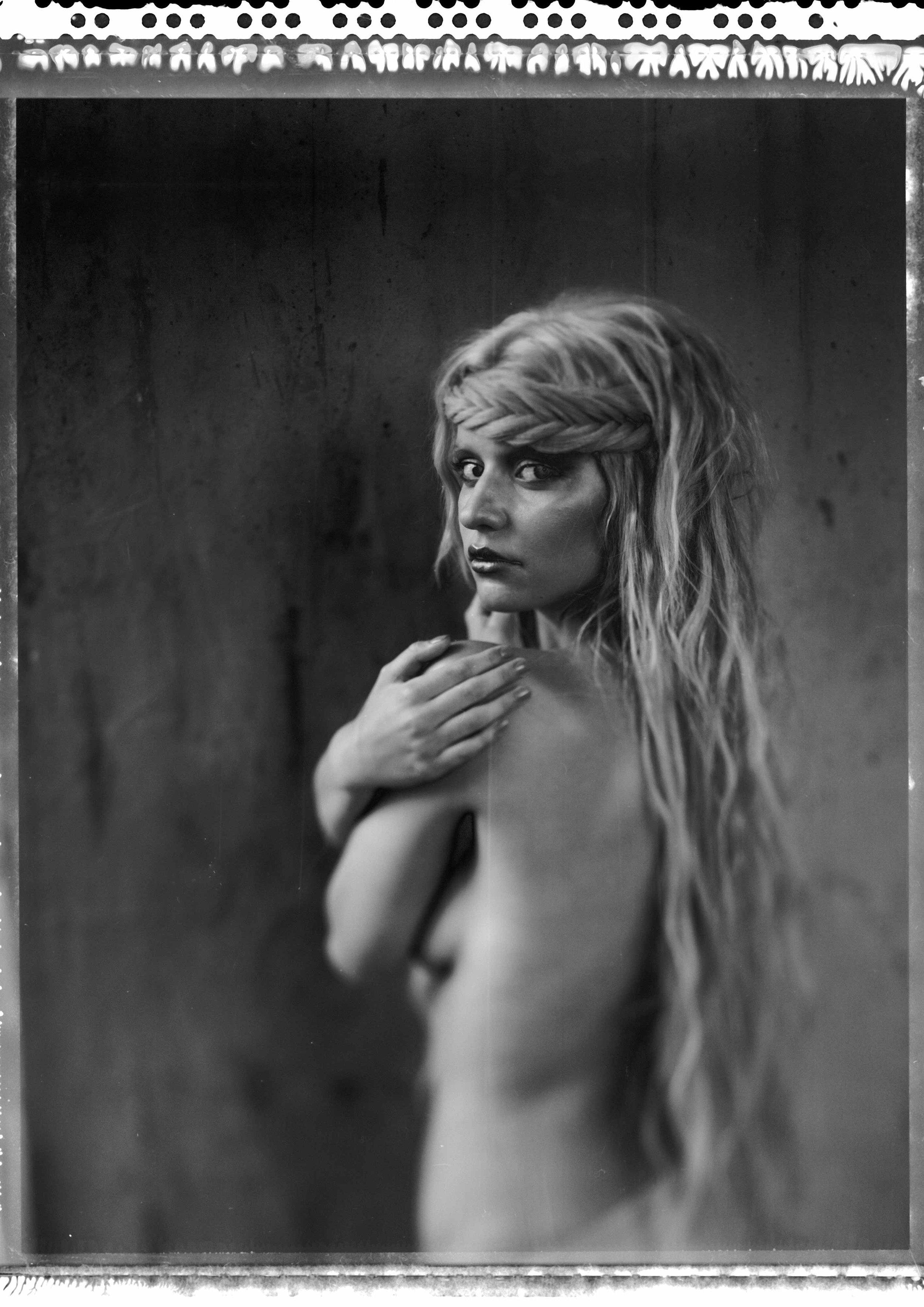 Schwarzweißfotografie einer Frau mit langem, welligem Haar und geflochtenem Haarband, die mit ernstem Ausdruck über die Schulter blickt, vor einem dunklen, strukturierten Hintergrund.
