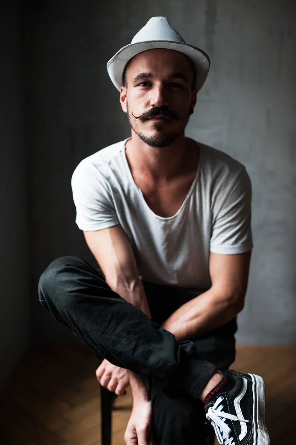 Mann mit Schnurrbart und Spitzbart, trägt weißen Fedora, graues T-Shirt, schwarze Hose und Sneakers, sitzt auf einem Stuhl vor einer grauen Wand.
