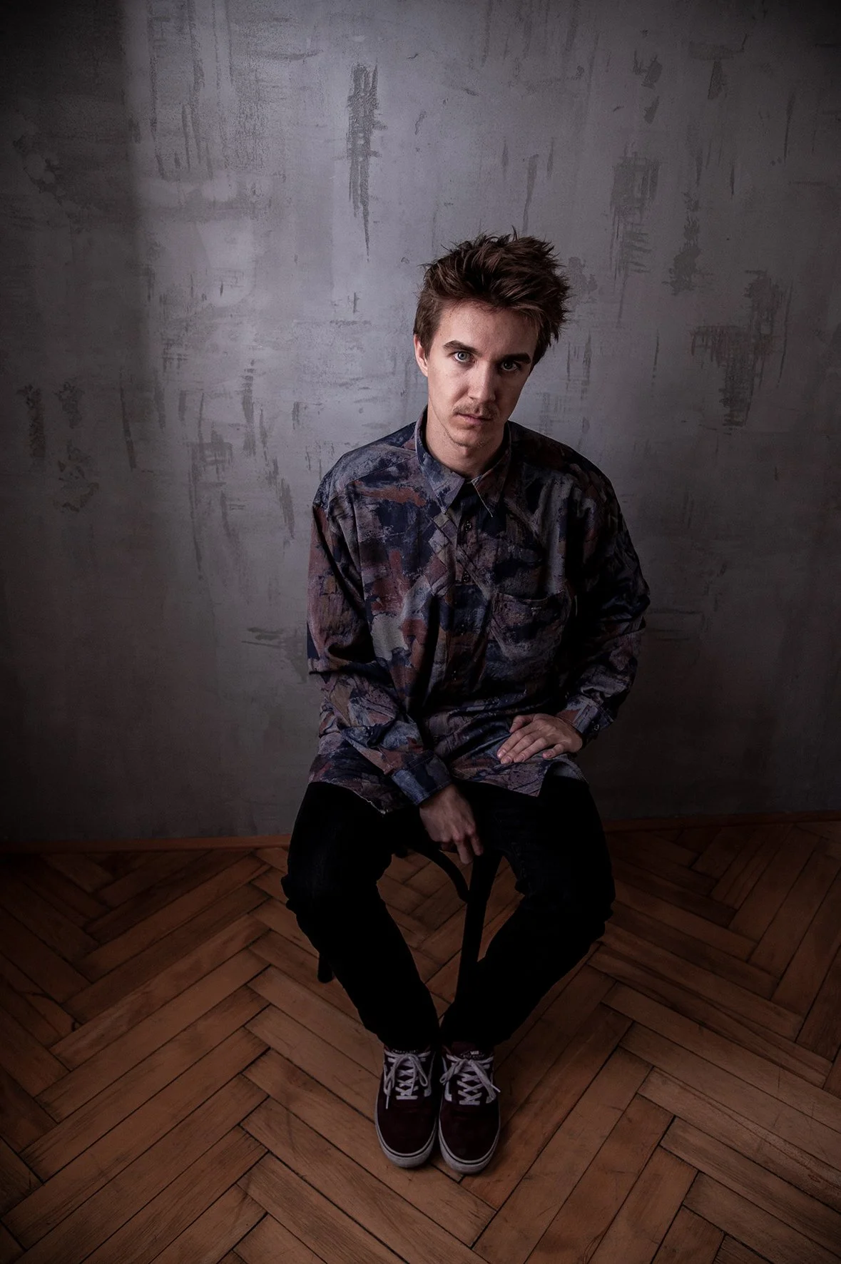 Ein junger Mann mit gestyltem braunem Haar sitzt auf einem schwarzen Stuhl, trägt ein mehrfarbig gemustertes Hemd mit Knopfleiste, schwarze Hose und dunkle Sneakers, in einem Studio mit strukturierter grauer Wand und Parkettboden.