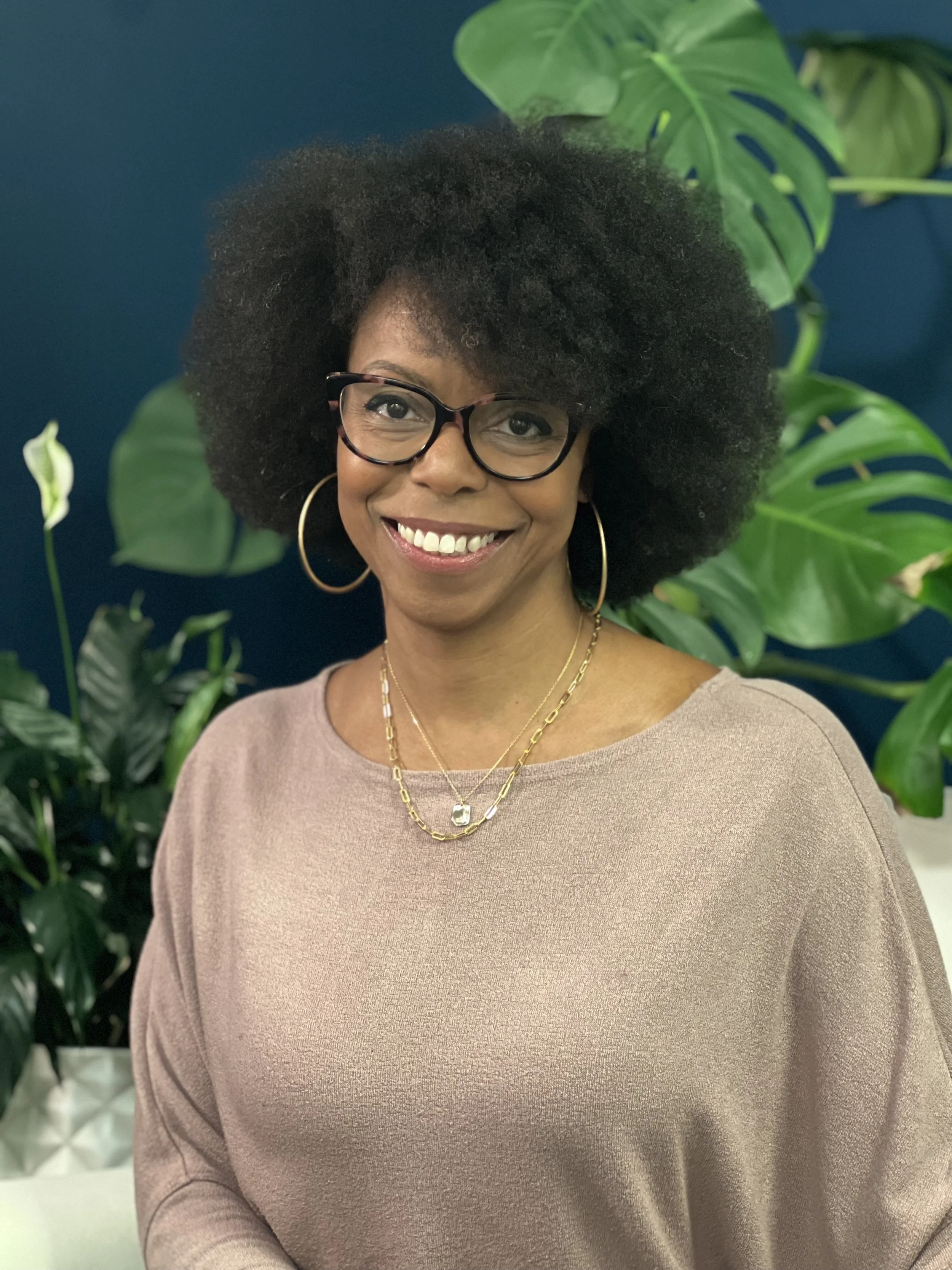 A woman with glasses and hoop earrings smiling in front of large green houseplants against a dark blue wall.