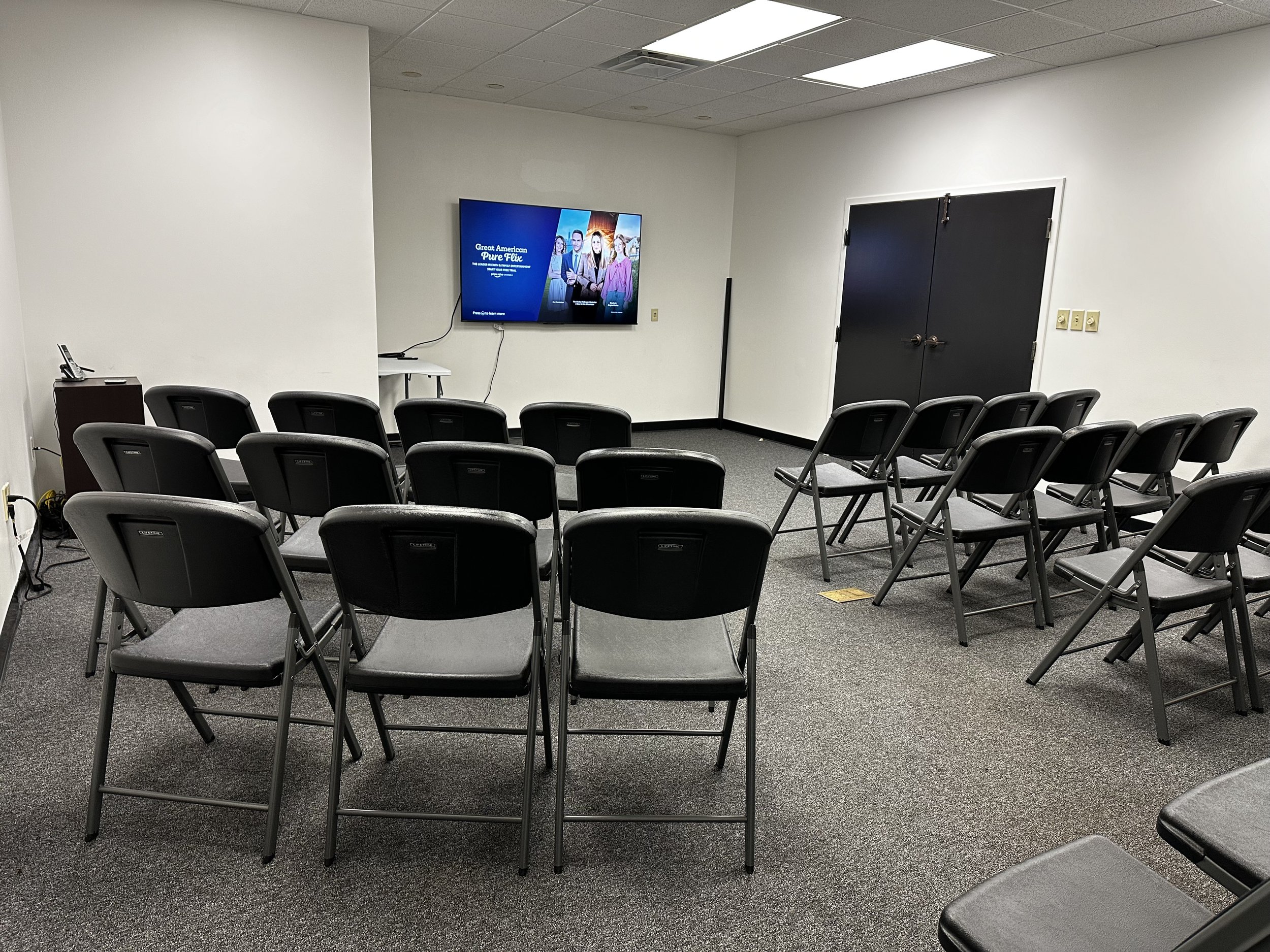 An empty conference or meeting room with black chairs arranged in rows facing a mounted television on a white wall. The room has a gray carpet, ceiling tiles with fluorescent lighting, and a closed double black door.