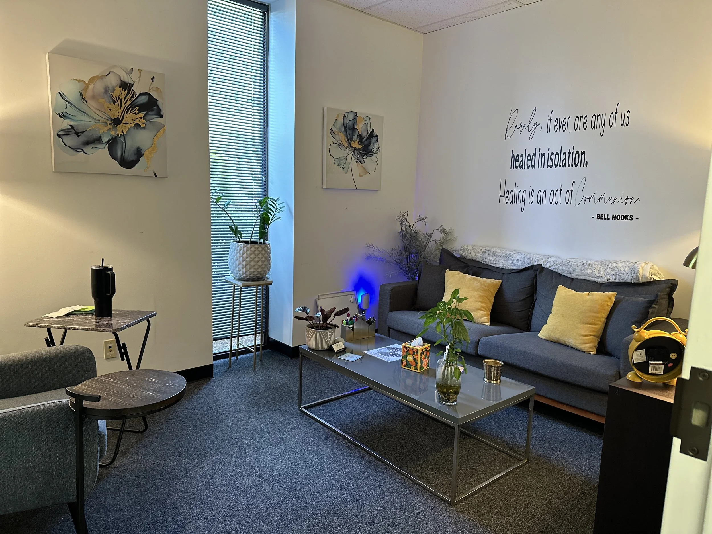 A cozy living room with a gray sofa with yellow and floral pillows, a glass coffee table with plants and decor, artwork of flowers on the walls, a window with vertical blinds, and a quote on the wall about healing and communication.