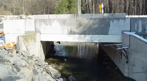 Concrete bridge over a creek with trees and a yellow road sign in the background.
