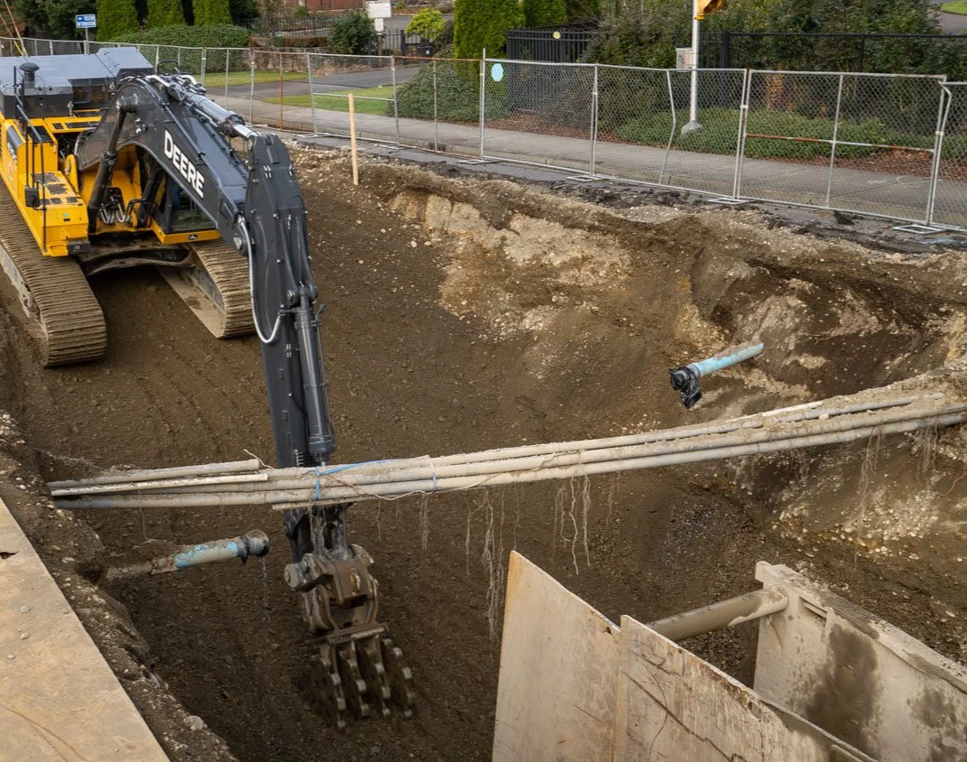 Construction equipment near a body of water under a bright sun, including a large orange excavator and a smaller piece of machinery, with construction tools and safety cones along the sidewalk.