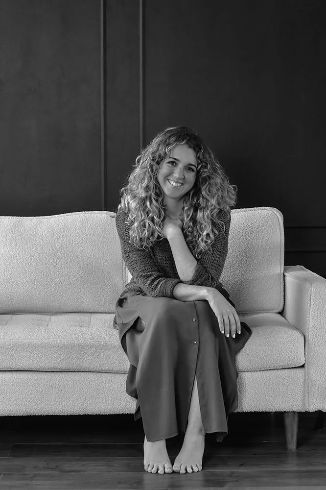 Woman with curly hair sitting on a sofa, smiling, with her legs crossed and her chin resting on her hand.