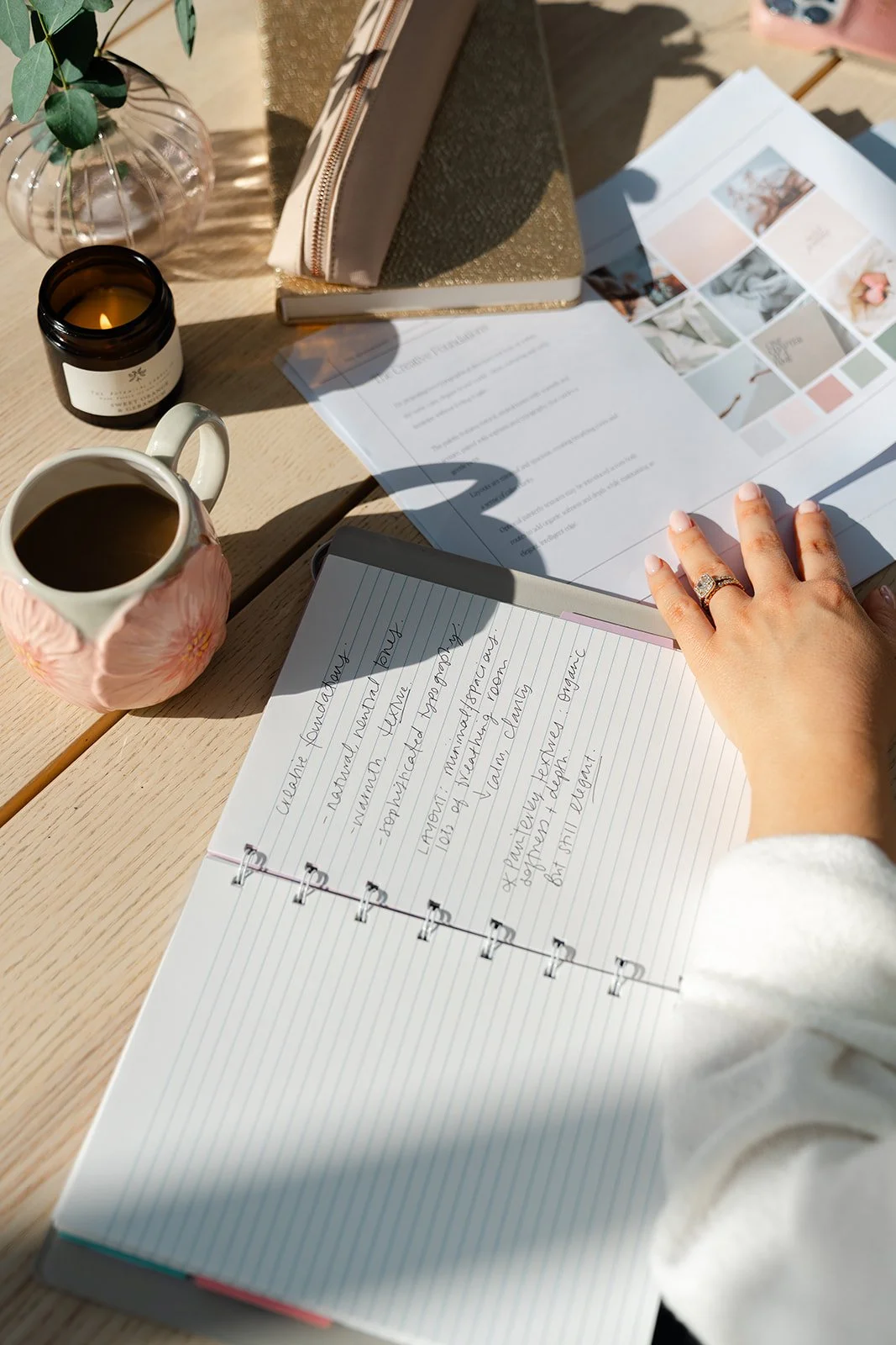 Overhead view of a wooden desk with a cup of coffee, a lit candle, a pink floral mug, a transparent vase with greenery, various notebooks, printed documents, and a person's hand with an engagement ring, all illuminated by natural sunlight.