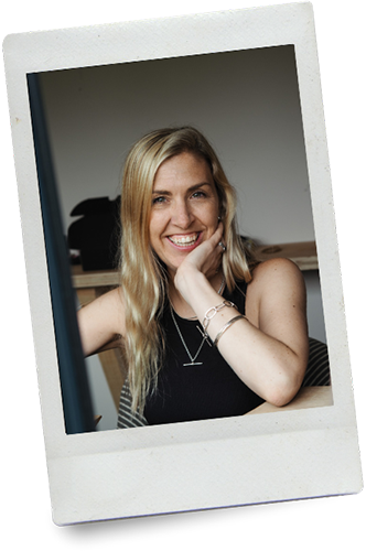 A young woman with blonde hair smiling and resting her chin on her hand, sitting at a desk in a home or office setting.
