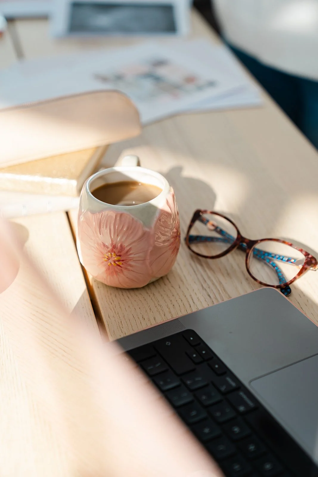 A desk with a ceramic mug of coffee, a pair of tortoiseshell glasses, an open book, and a laptop.