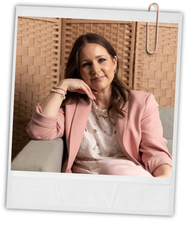 A woman with long brown hair sits on a light gray couch, resting her chin on her hand, wearing a pink blazer and a white lace top, in front of a wooden woven backdrop, with a paper clip attached at the top of the image.