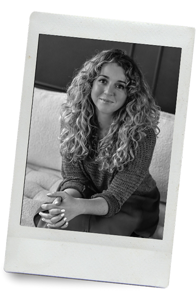 A young woman with curly hair sitting on a couch, smiling at the camera, in black and white.
