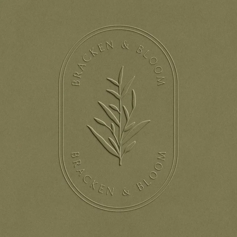 Embossed logo with an illustration of a branch and the text 'Bracken & Bloom' enclosed in an oval outline on a green background.