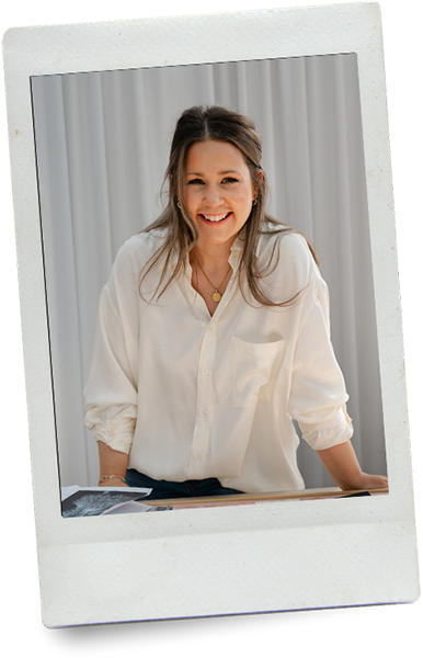 Brand and Website designer with long hair smiling, wearing a white shirt, leaning on a table with a gray curtain background.