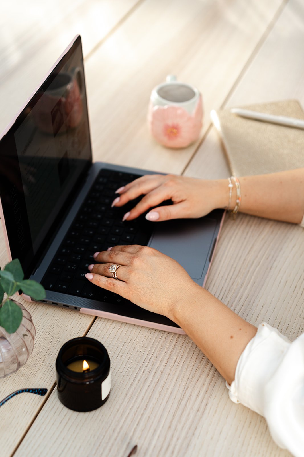 A brand and website designer  typing on a laptop with a potted plant, a candle, a pink mug, a pen, and a notebook on a wooden table.
