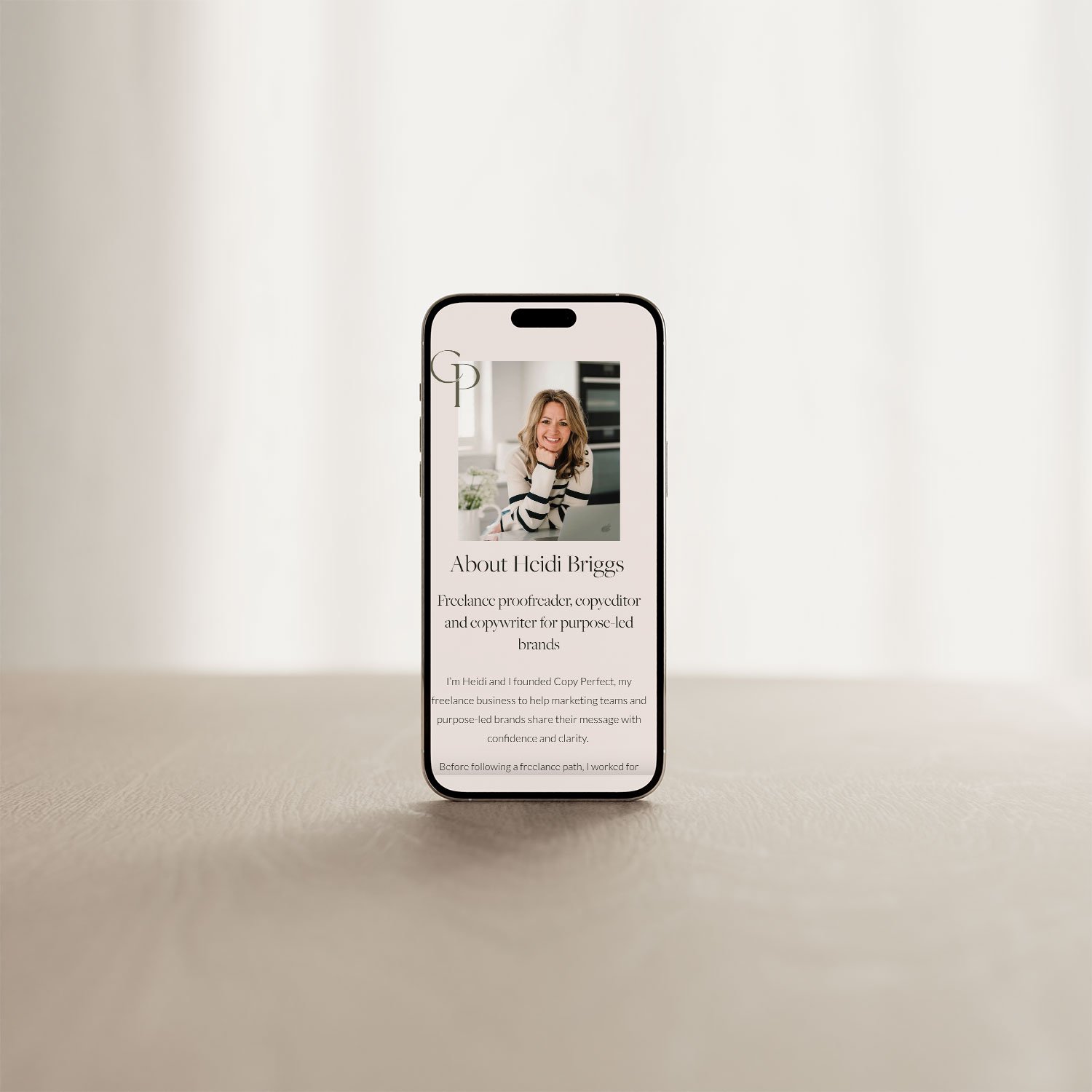 A smartphone displaying a profile page of a woman named Heidi Briggs, featuring her photo, name, and a brief biography about her freelance proofreading, copyediting, and copywriting services, set against a plain neutral background.