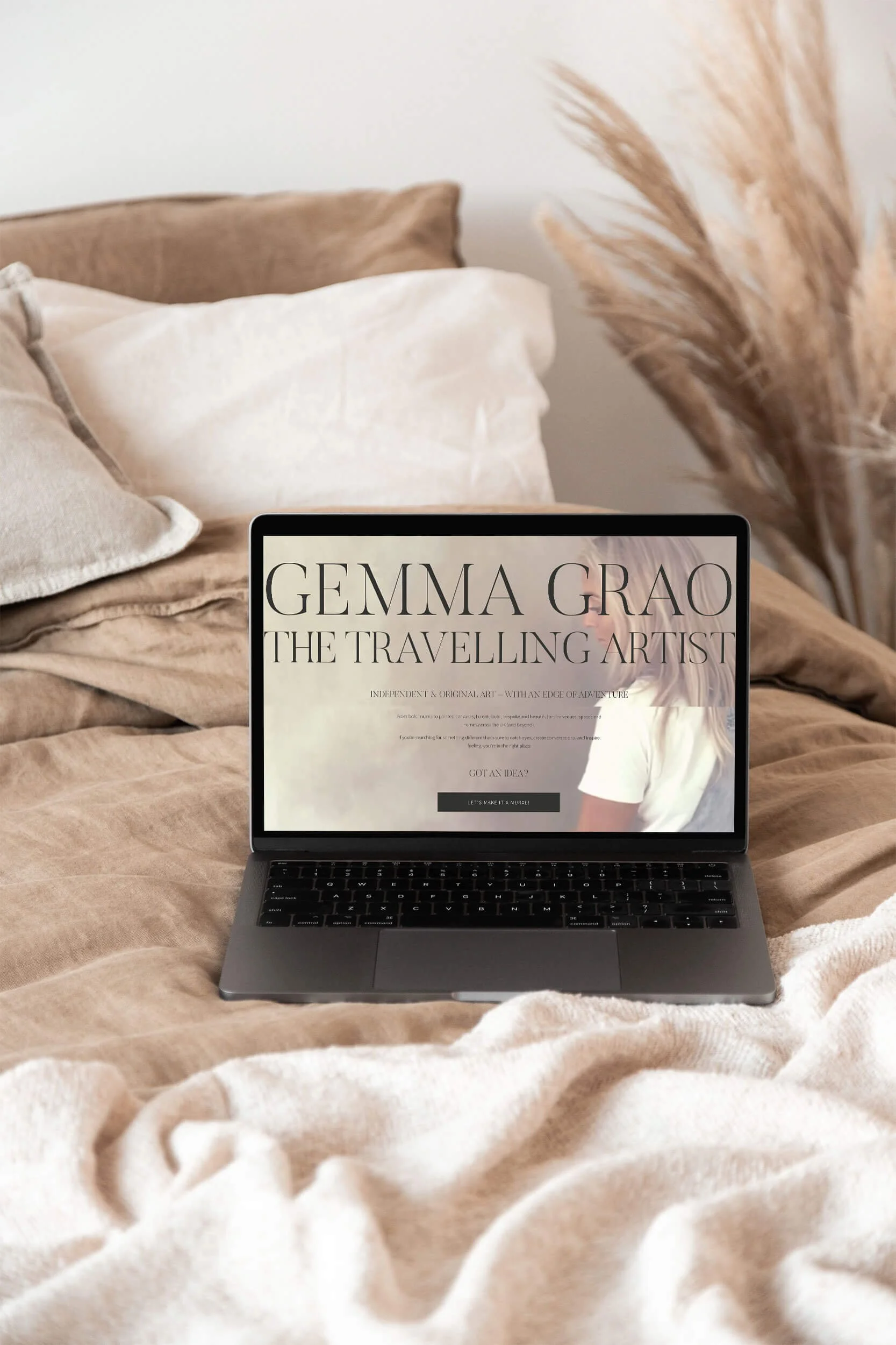 A laptop on a bed displaying a website about Gemma Grao, the traveling artist, with beige and white bedding and dried pampas grass in the background.