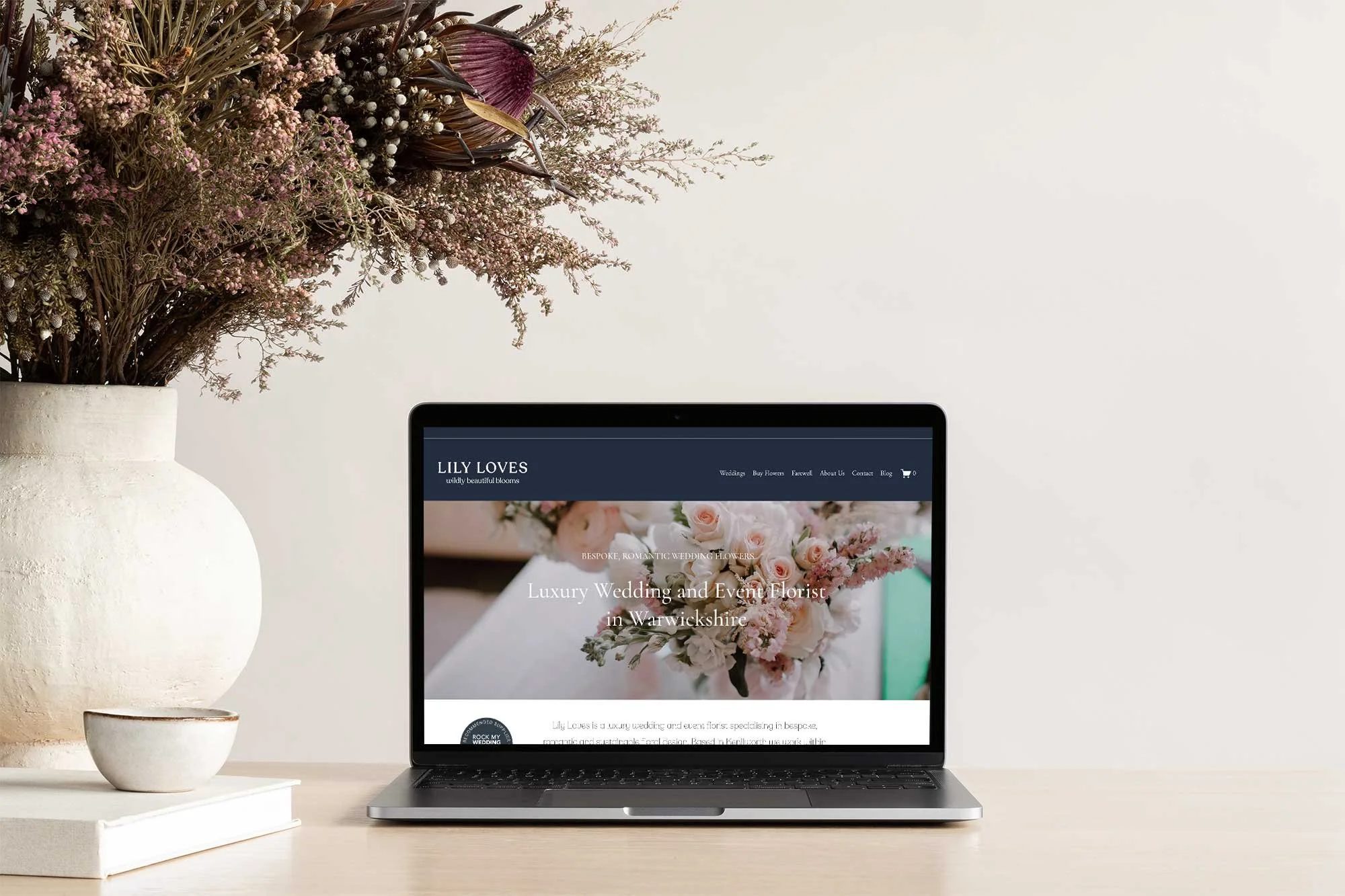 A computer screen displaying a wedding floristry website with a bouquet of pink and white flowers, alongside a large floral arrangement in a white vase on a light-colored desk, with a neutral wall in the background.