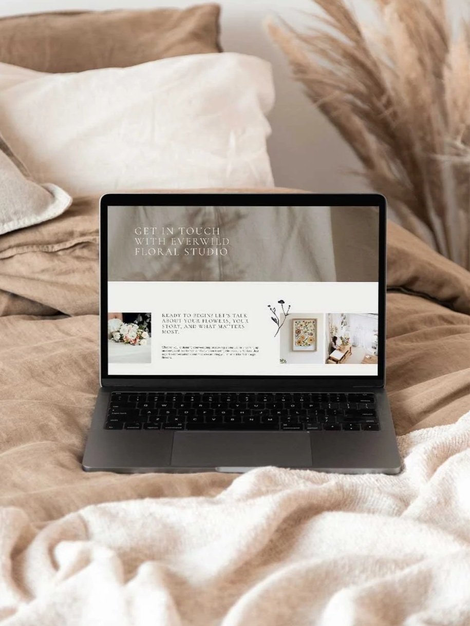 Open laptop on bed displays a floral studio website, surrounded by beige blankets and pillows in a cozy bedroom.