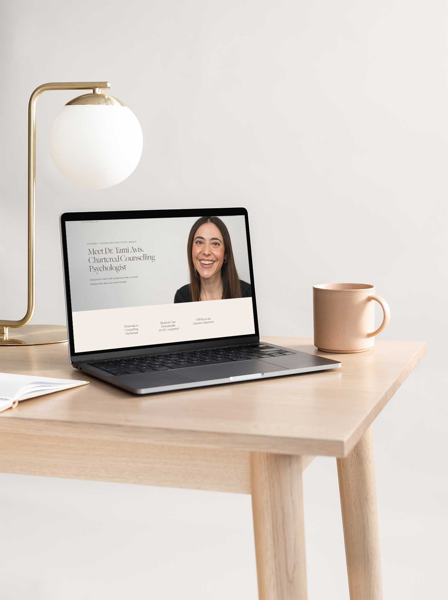 A wooden desk with a beige mug, an open notebook, a laptop displaying a woman and text about Dr. Tami Avis, a gold desk lamp with a white round shade, and a white wall in the background.