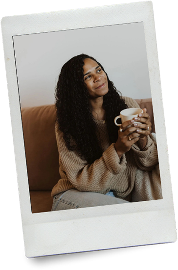 A woman sitting on a couch holding a white mug, looking thoughtfully to the side.
