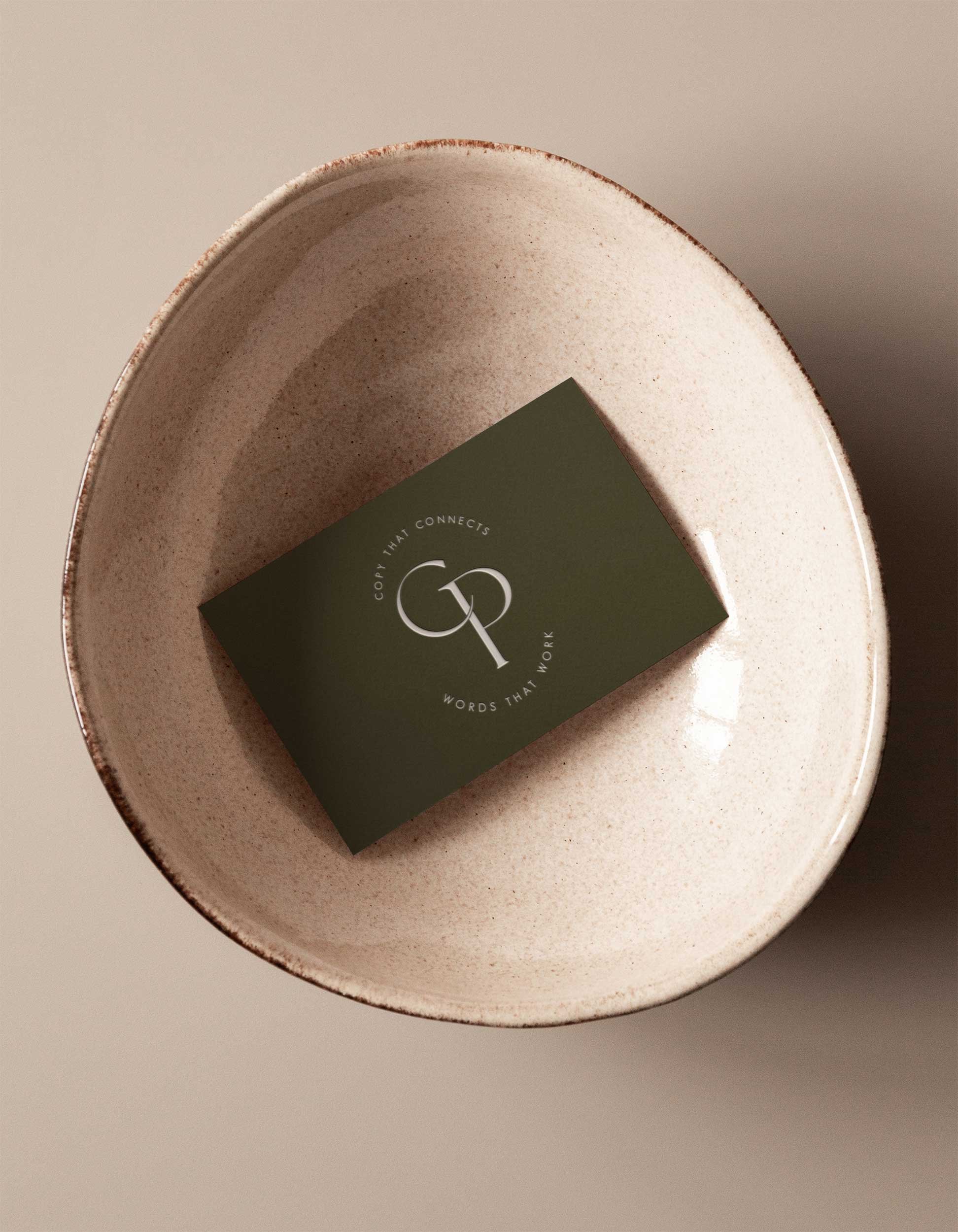 Close-up of an oval ceramic dish with a speckled beige interior and a slightly irregular rim, holding a green card or paper with white and gold text.