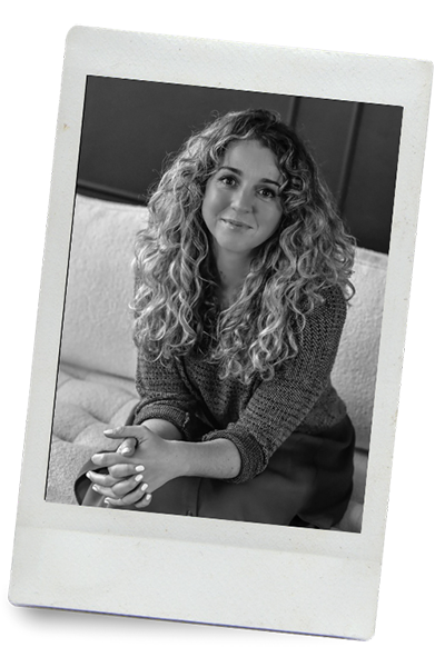 Black and white photo of a woman with curly hair, sitting on a couch and smiling at the camera.