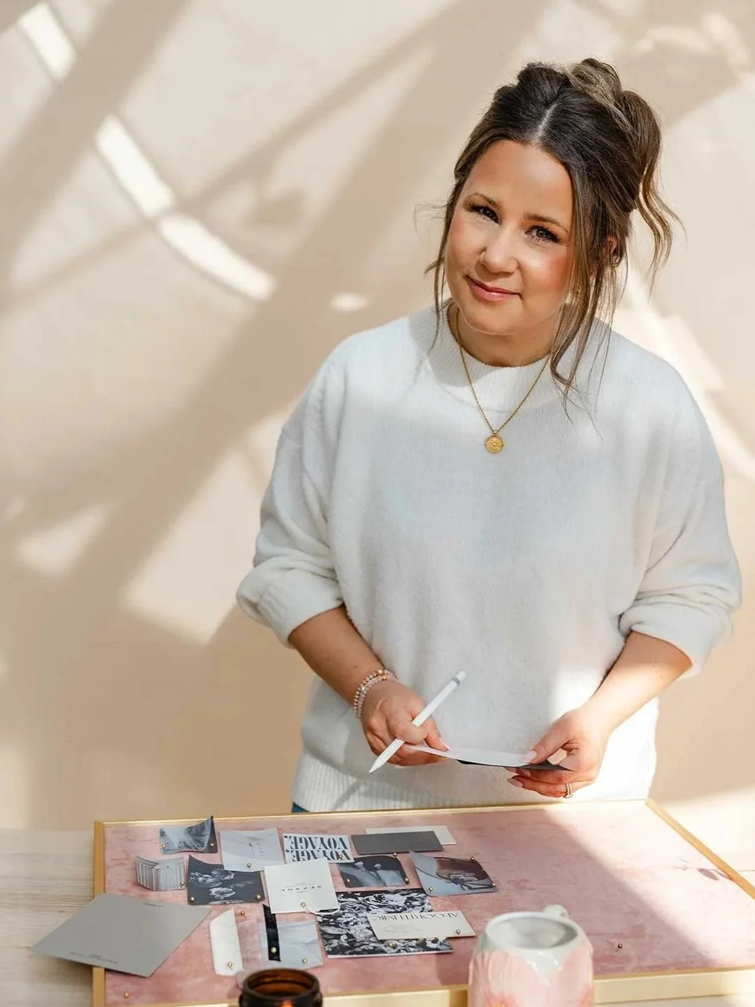 Brand and Website designer with brown hair and a white sweater, holding a white stylus and paper, standing at a table with photographs and items, in a softly lit room.