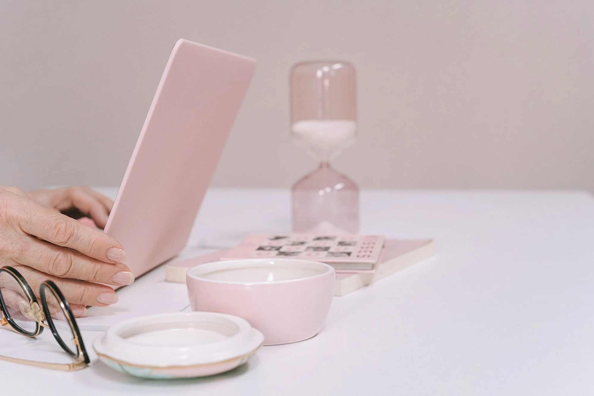 Close-up of a person's hand resting on a white surface, with pink cups, a pair of eyeglasses, a pink photo album, a pink and clear hourglass, and a notepad with black printed photos on it.