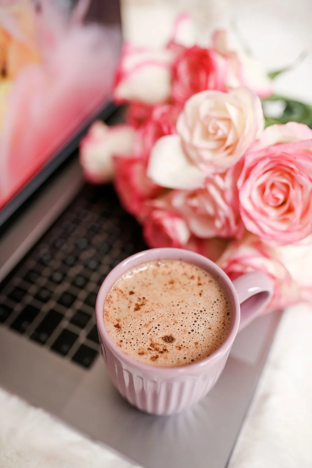 A pink mug of frothy coffee with cinnamon, a bouquet of pink and peach roses, and a laptop on a white surface.