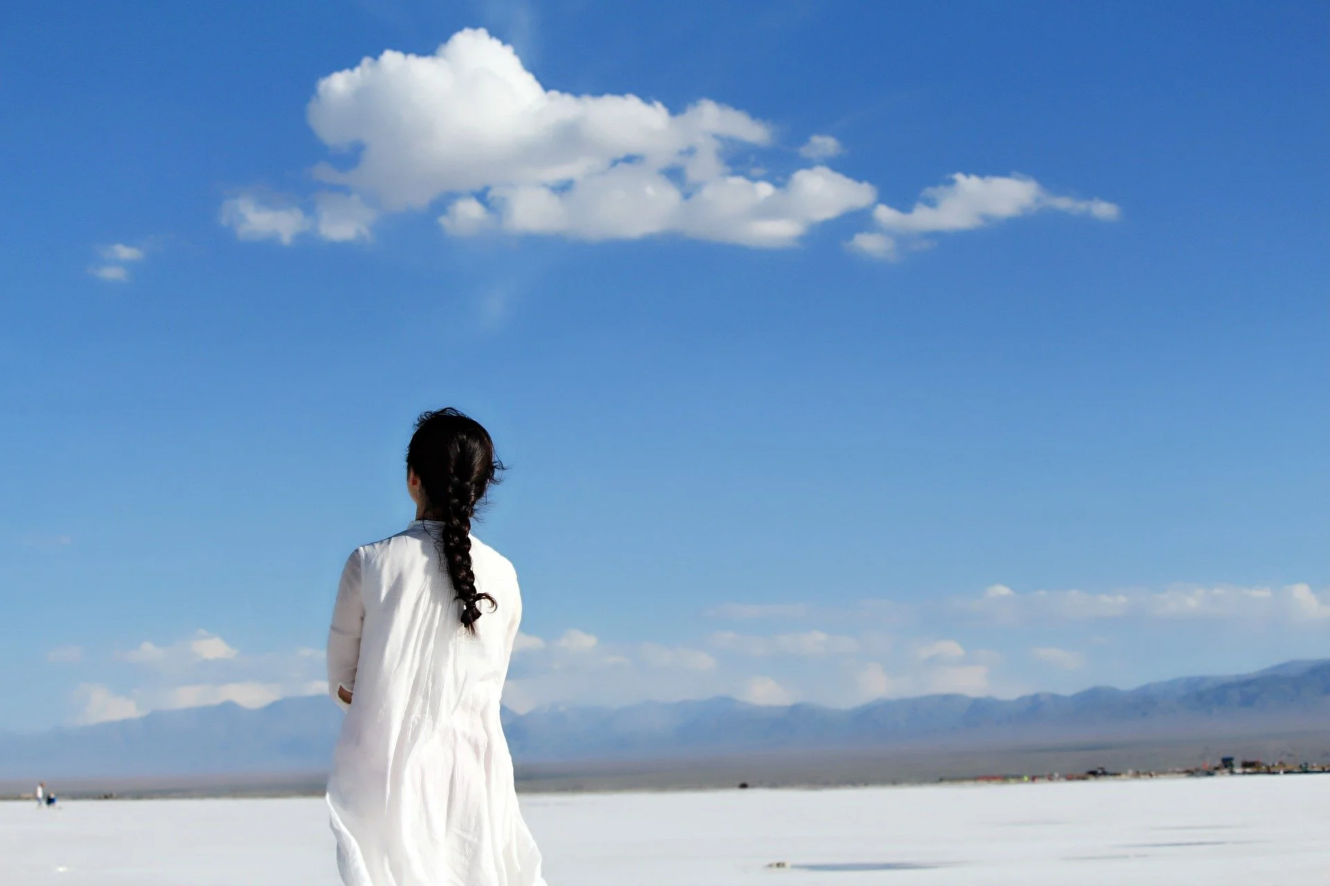 A woman with long braided hair wearing a white dress standing on a vast, flat landscape with distant mountains and a blue sky with scattered clouds.