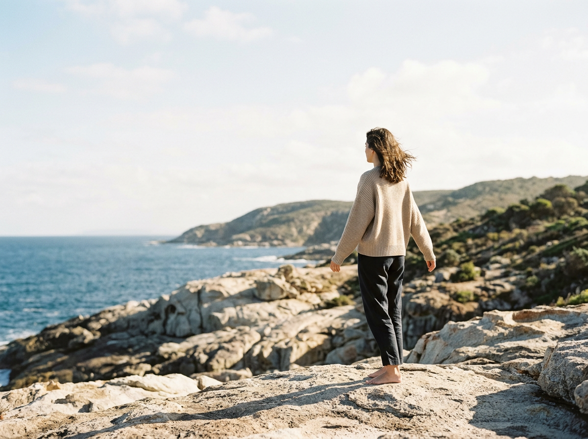 A woman standing barefoot on rocky terrain by the ocean, looking towards the horizon with hills in the background on a partly cloudy day.