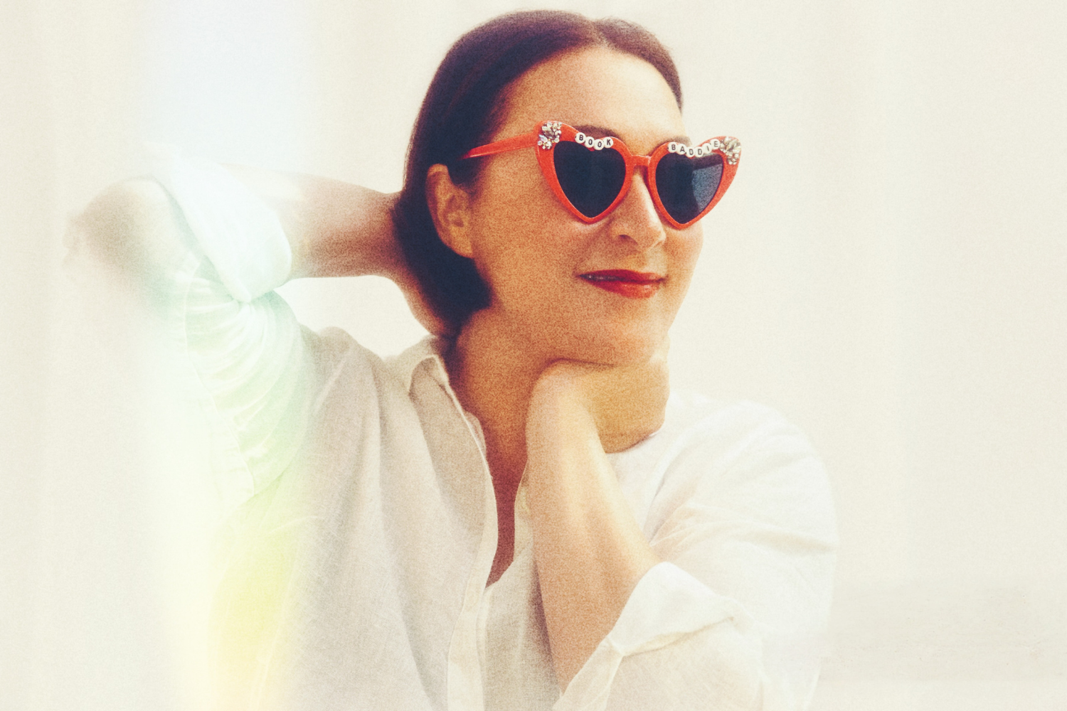 Woman with short brown hair wearing heart-shaped sunglasses with 'Book Baddie' written on the frames, smiling, with her hand resting on her neck and chin, against a plain light background.