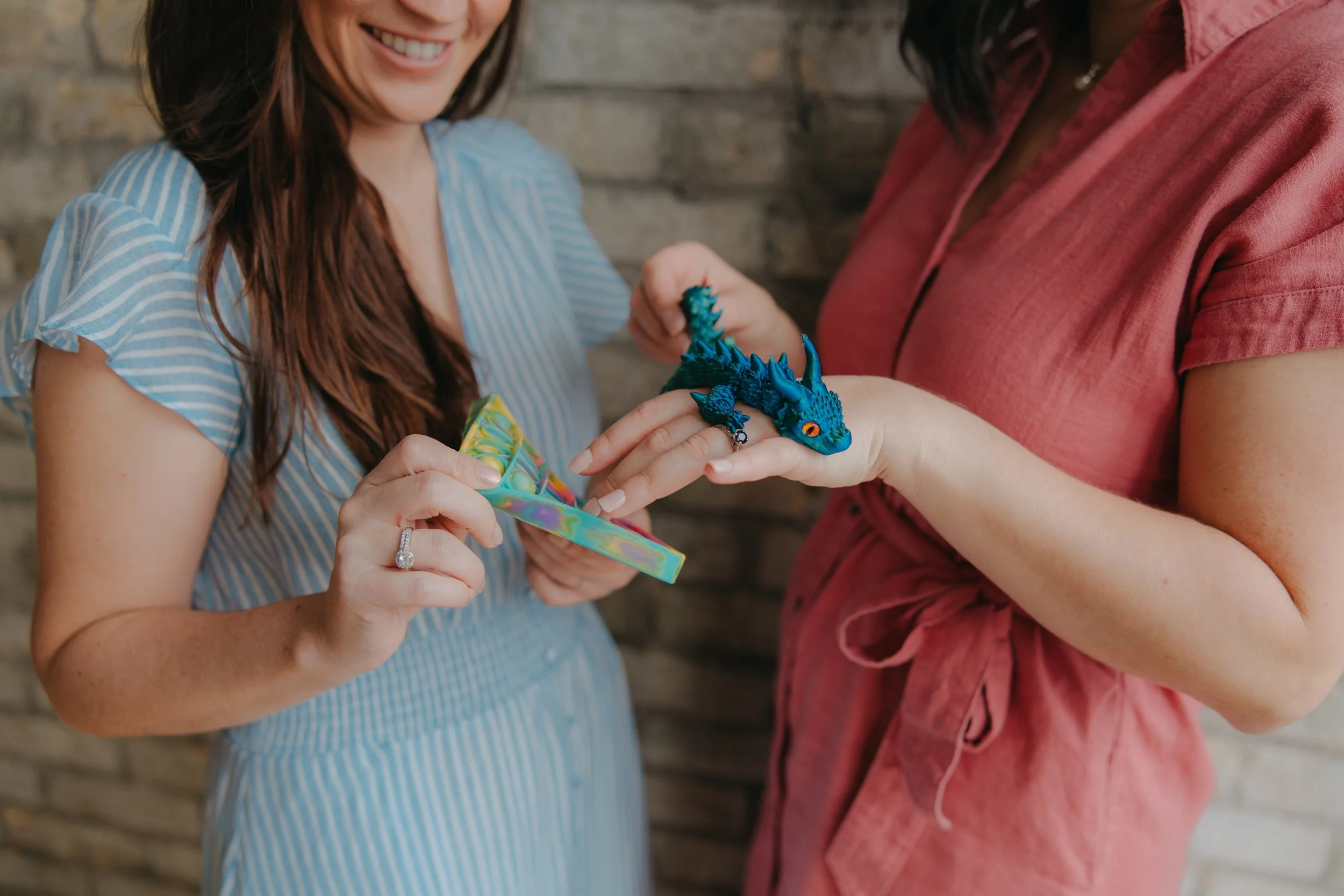 Two women, one wearing a blue and white striped dress and the other a red dress, handling a small blue dragon plush toy and a colorful container, with a brick wall in the background.