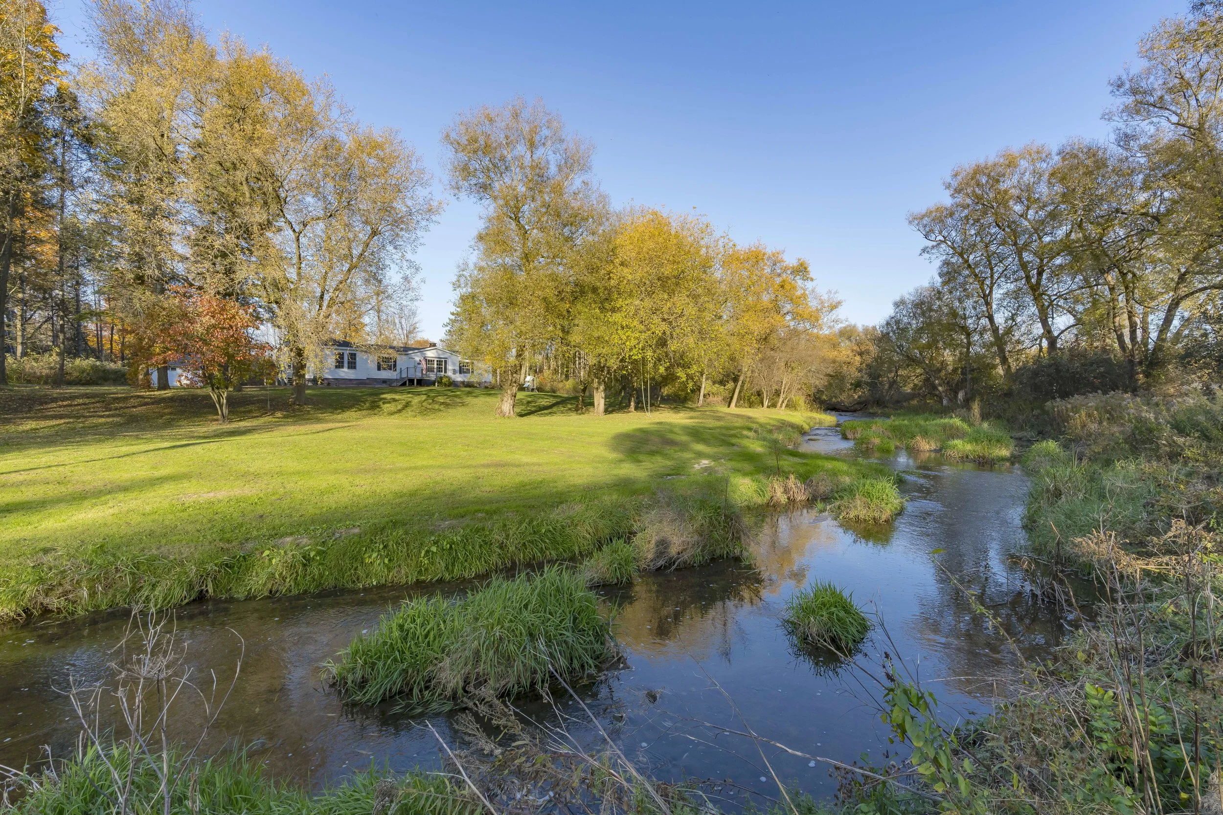 A scenic landscape with a small stream flowing through lush green grass and trees with some leaves changing color, with a house in the background under a clear blue sky.