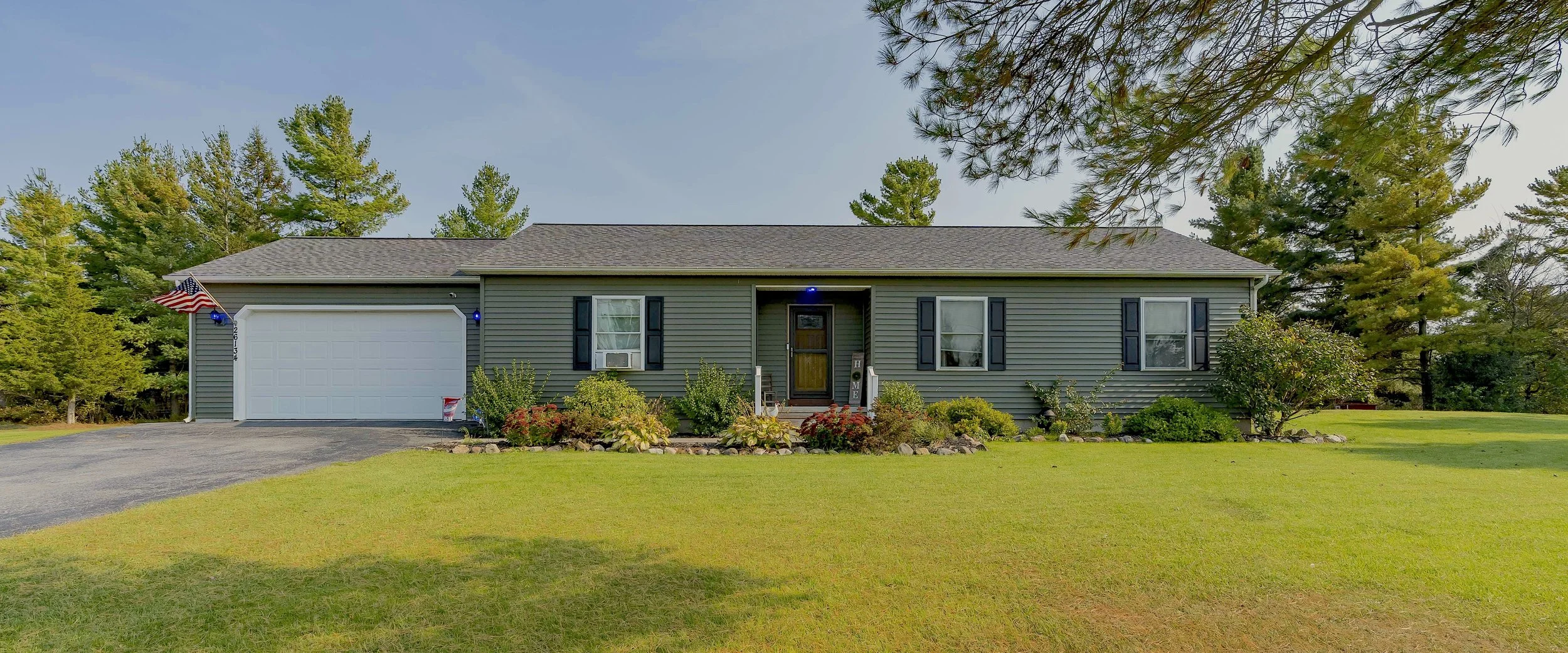 A single-story gray house with a brown door, white garage, and black shutters, surrounded by a green lawn and trees, with an American flag hanging on the left side.