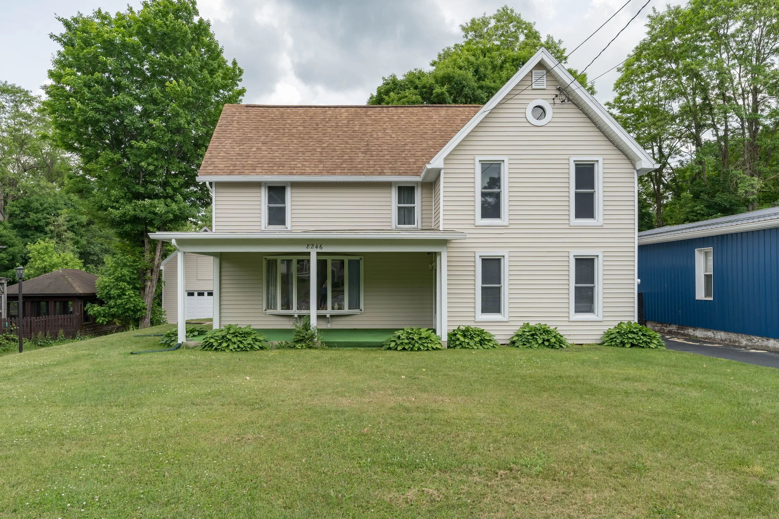 A two-story house with beige siding, a gable roof, and a porch with white columns. There are six tall, narrow windows on the front, a small round window near the top of the gable, and lush greenery in the yard.