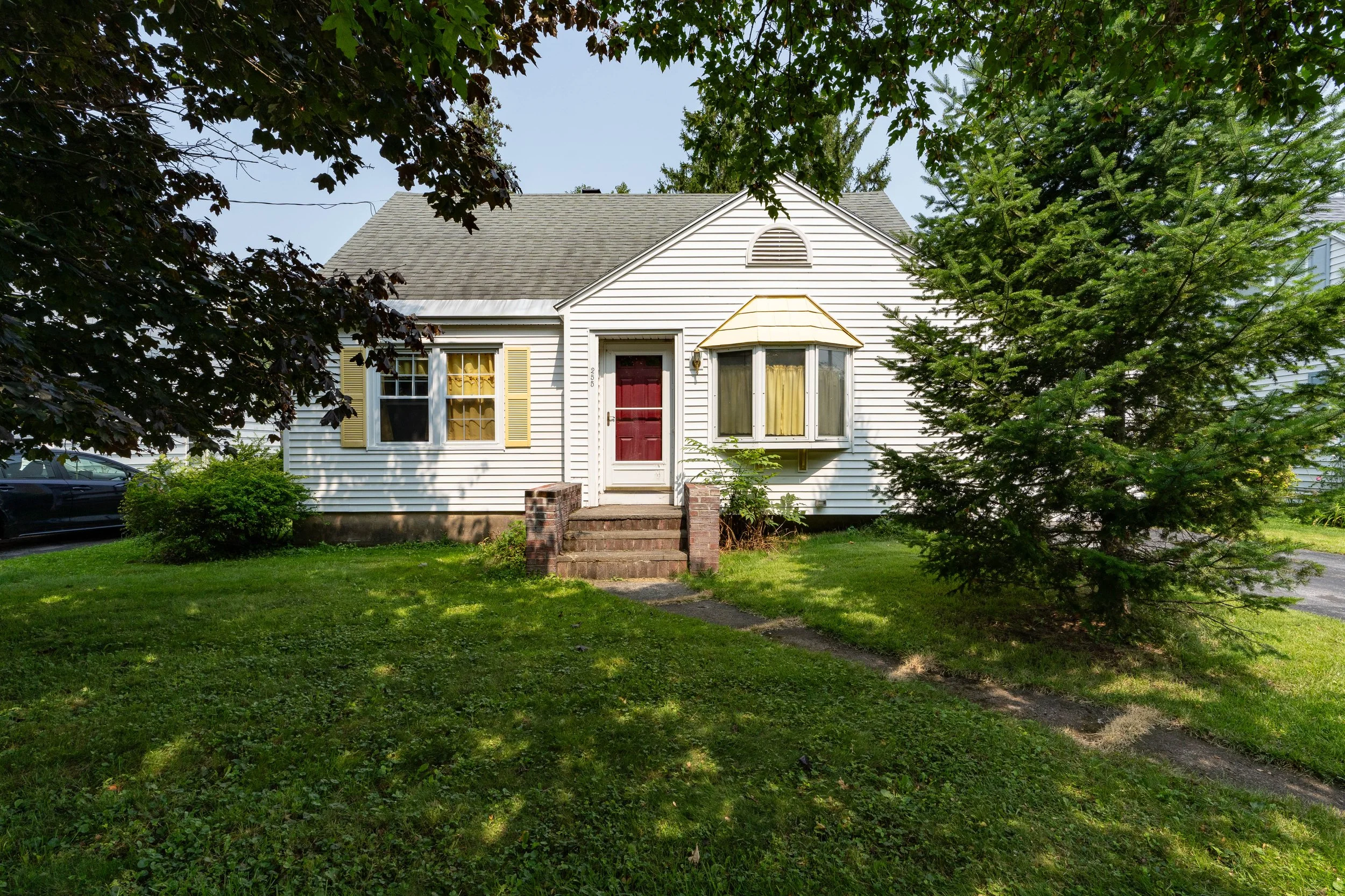 A small white house with yellow shutters, a red door, dormer window, and a bay window on the front, surrounded by green grass and trees.