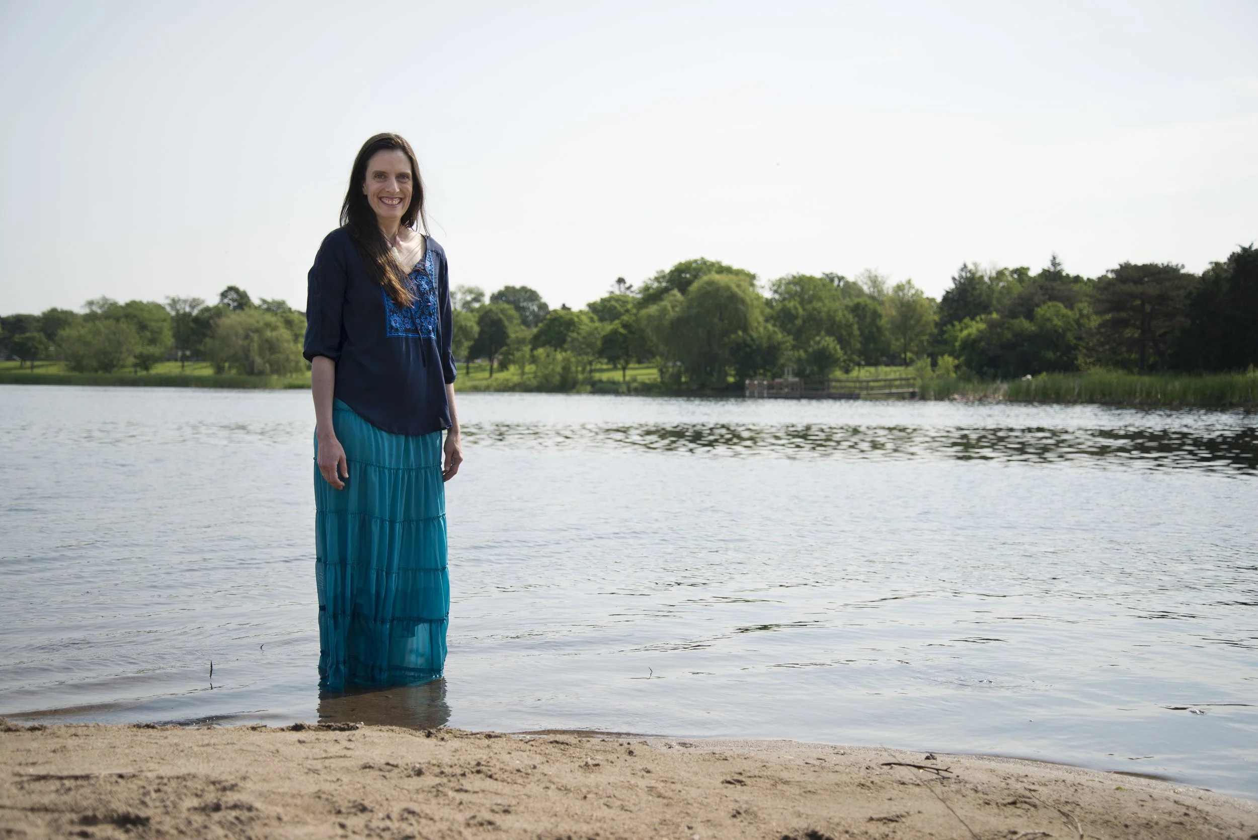 Woman standing in shallow water at lakeside, smiling, wearing a blue blouse and matching long skirt, with trees and grassy shoreline in the background on a clear day.